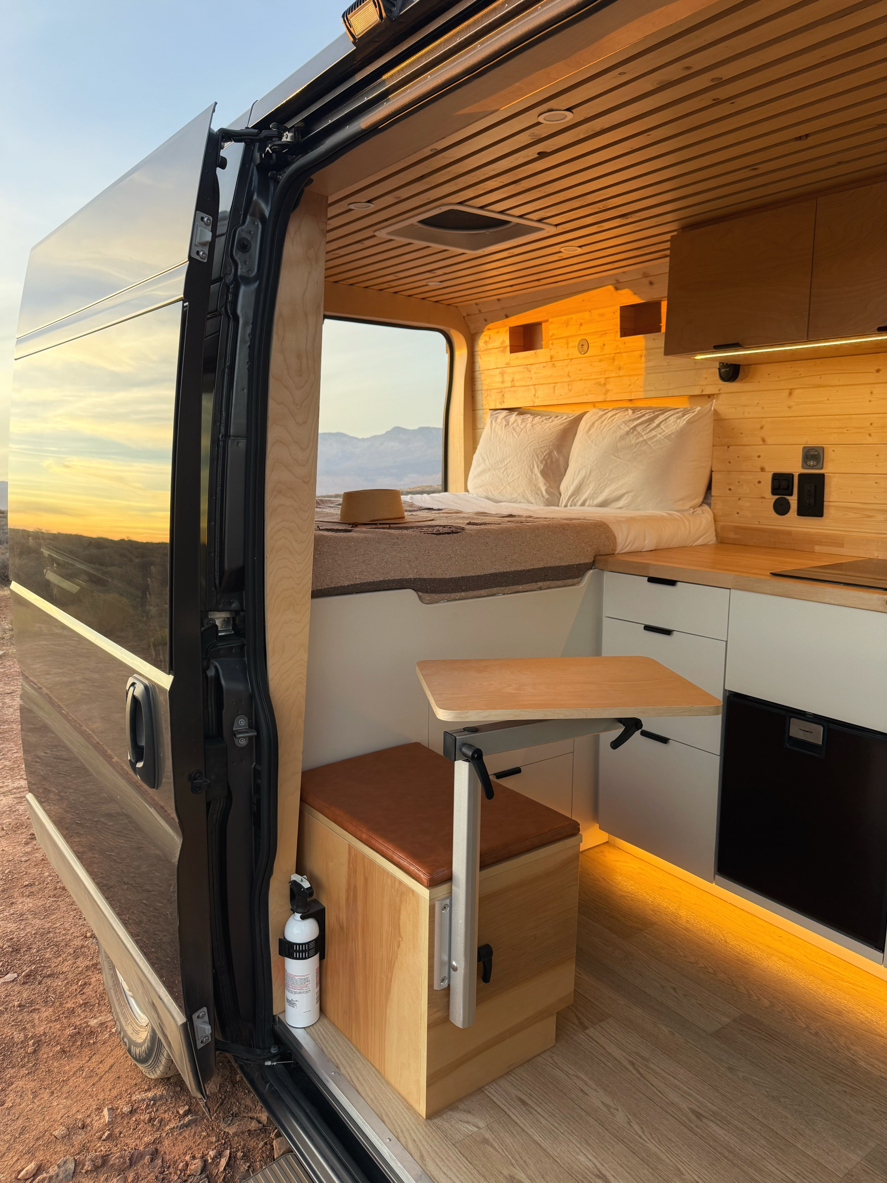 Inside an RV rental: a modern camper van with cozy wood interior, bed, kitchenette, and a flexible dining table, featuring a sunset view.. Custom RAM Promaster 2023