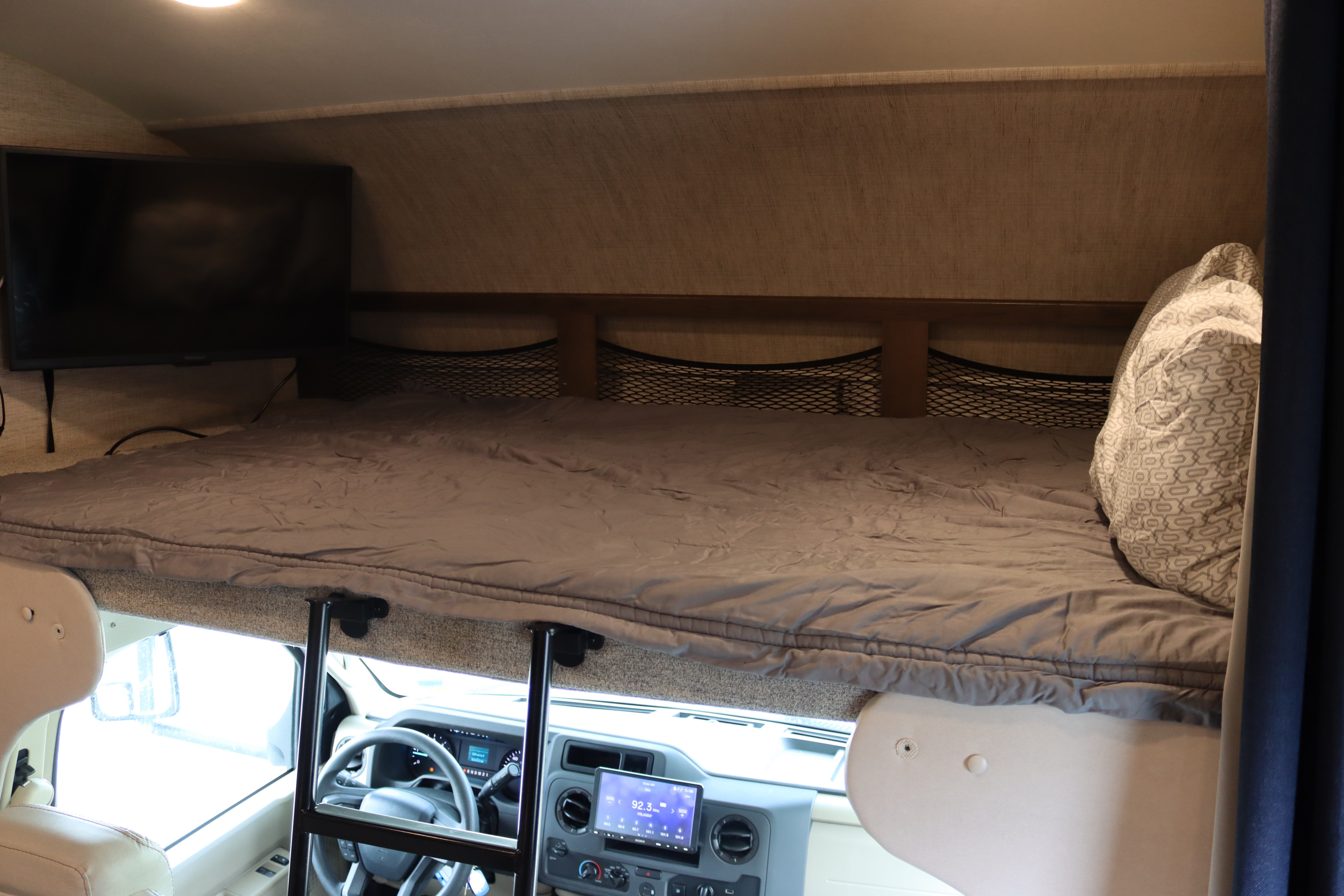 RV rental's cab-over bunk bed with grey bedding and TV. Below is the driver's area with steering wheel and dashboard.. Jayco Redhawk 2021
