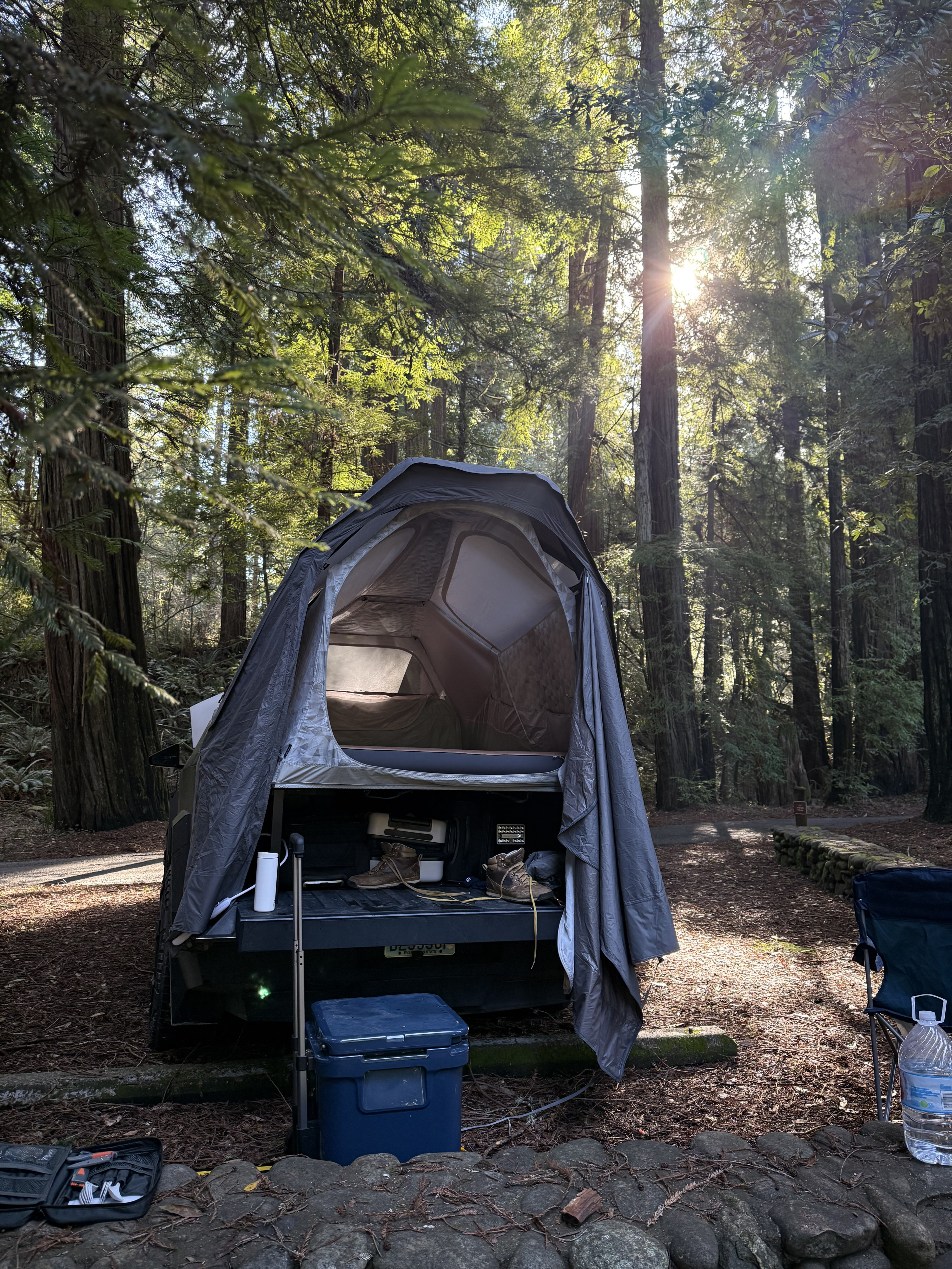 Truck bed tent camping setup in a sunlit forest, offering an adaptable RV rental alternative for adventures.. Tesla Cybertruck 2024