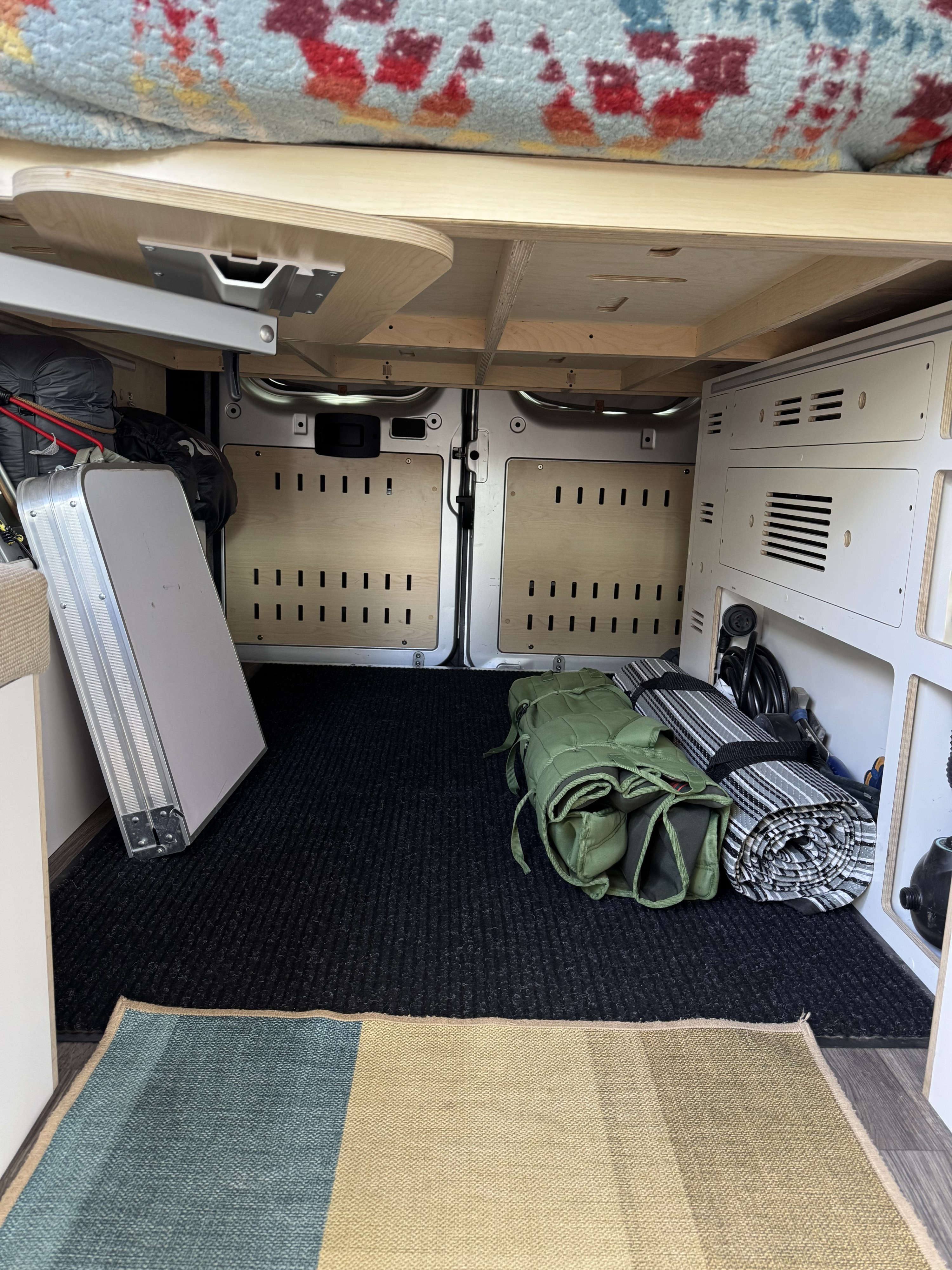 Organized under-bed storage in an RV rental, featuring a stowed table, camping gear, and rear van door paneling.. Dave and Matt Vans LV7 Ram Promaster 3500 2022