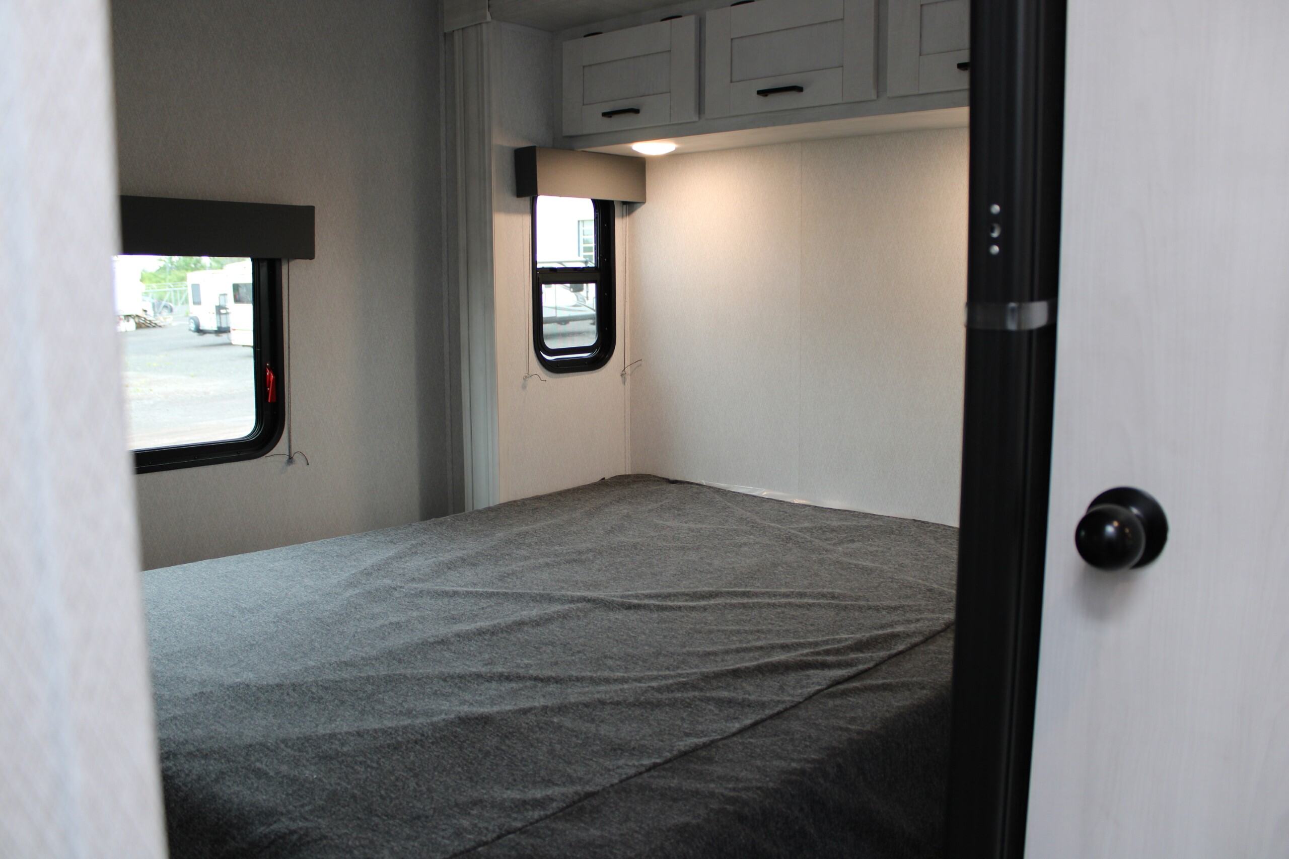 Inside an RV rental, showing a neatly made bed, two windows for natural light, and ample overhead cabinets.. EAST TO WEST ENTRADA 2023
