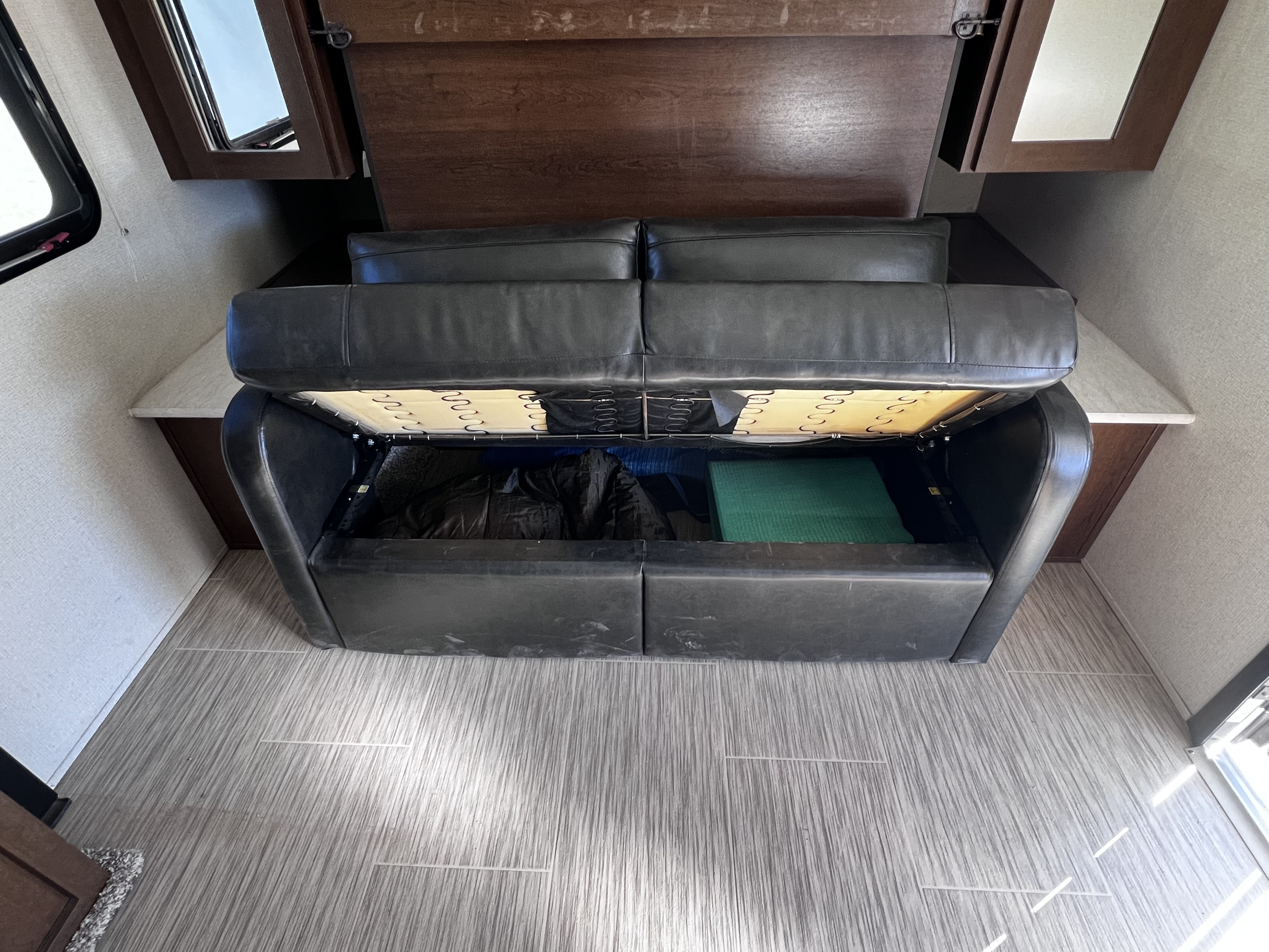 RV rental sofa bed with lifted cushion, revealing under-seat storage containing a black bag and green mat.. Forest River Tracer Breeze 2018
