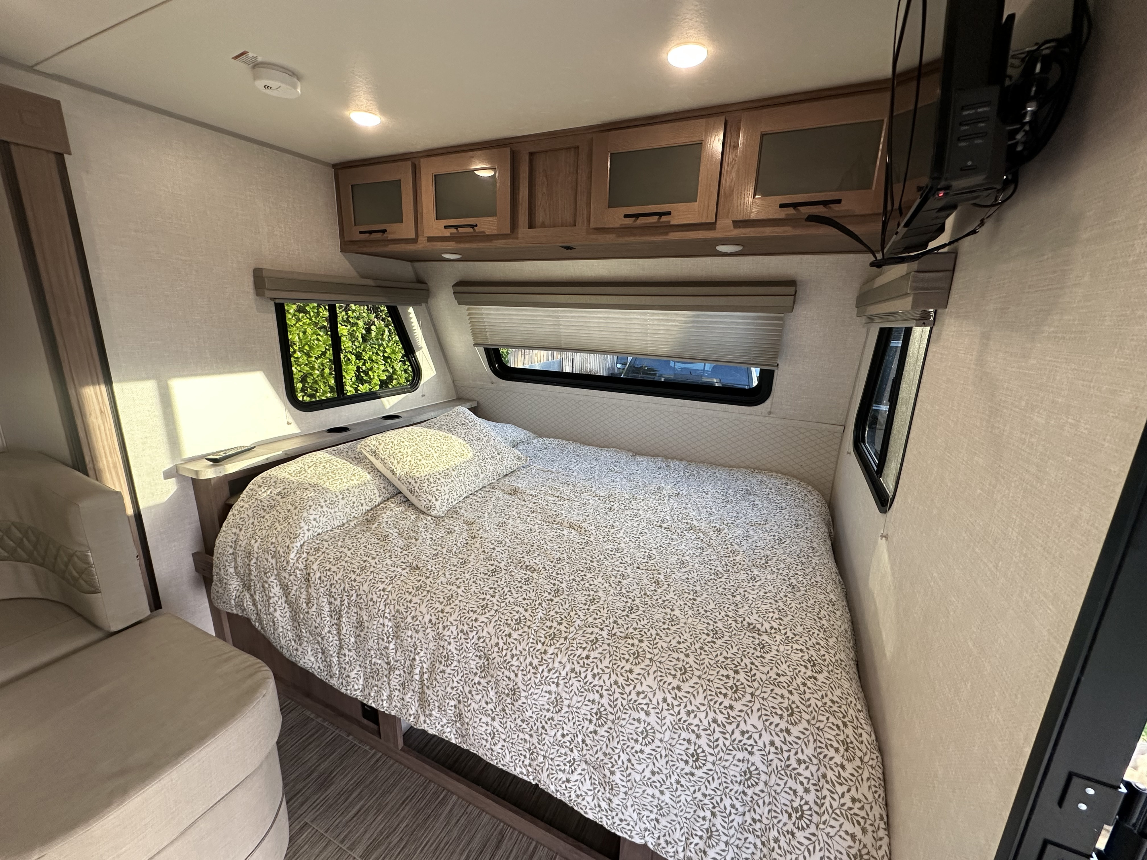 Bright RV rental bedroom with patterned bed, overhead storage, wall-mounted TV, and cozy window views.. No Boundaries 19 2021