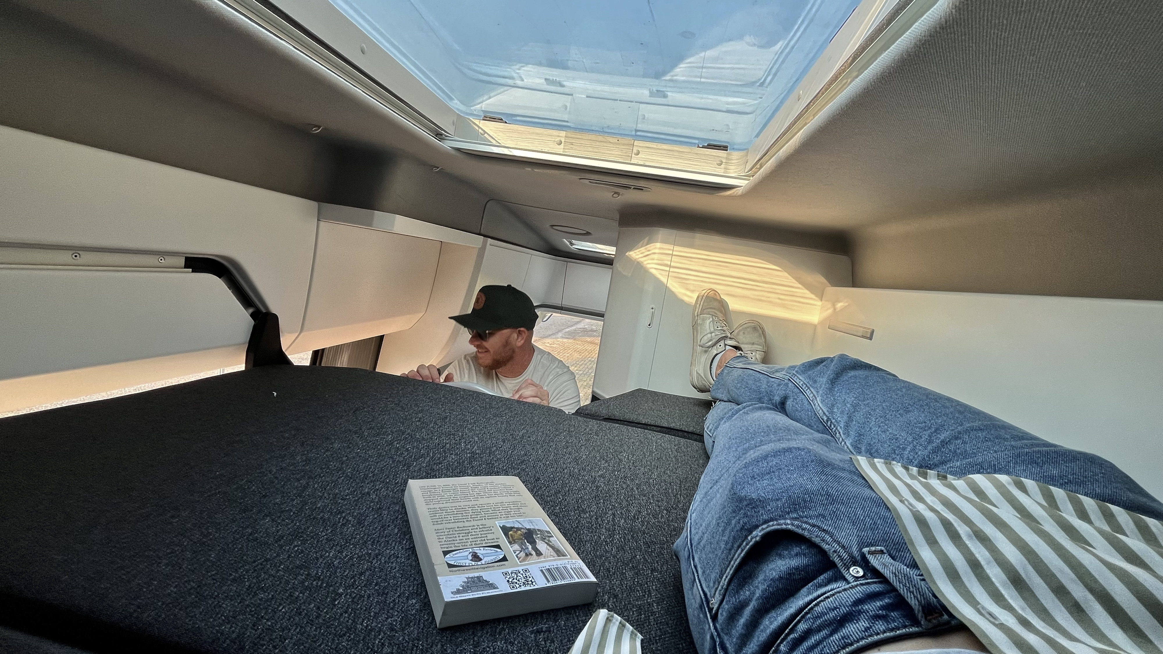 Relaxing in an RV rental with legs up and a book, as a man peers in from the front. Bright skylight above.. Volkswagen Grand California - Clyde 2022