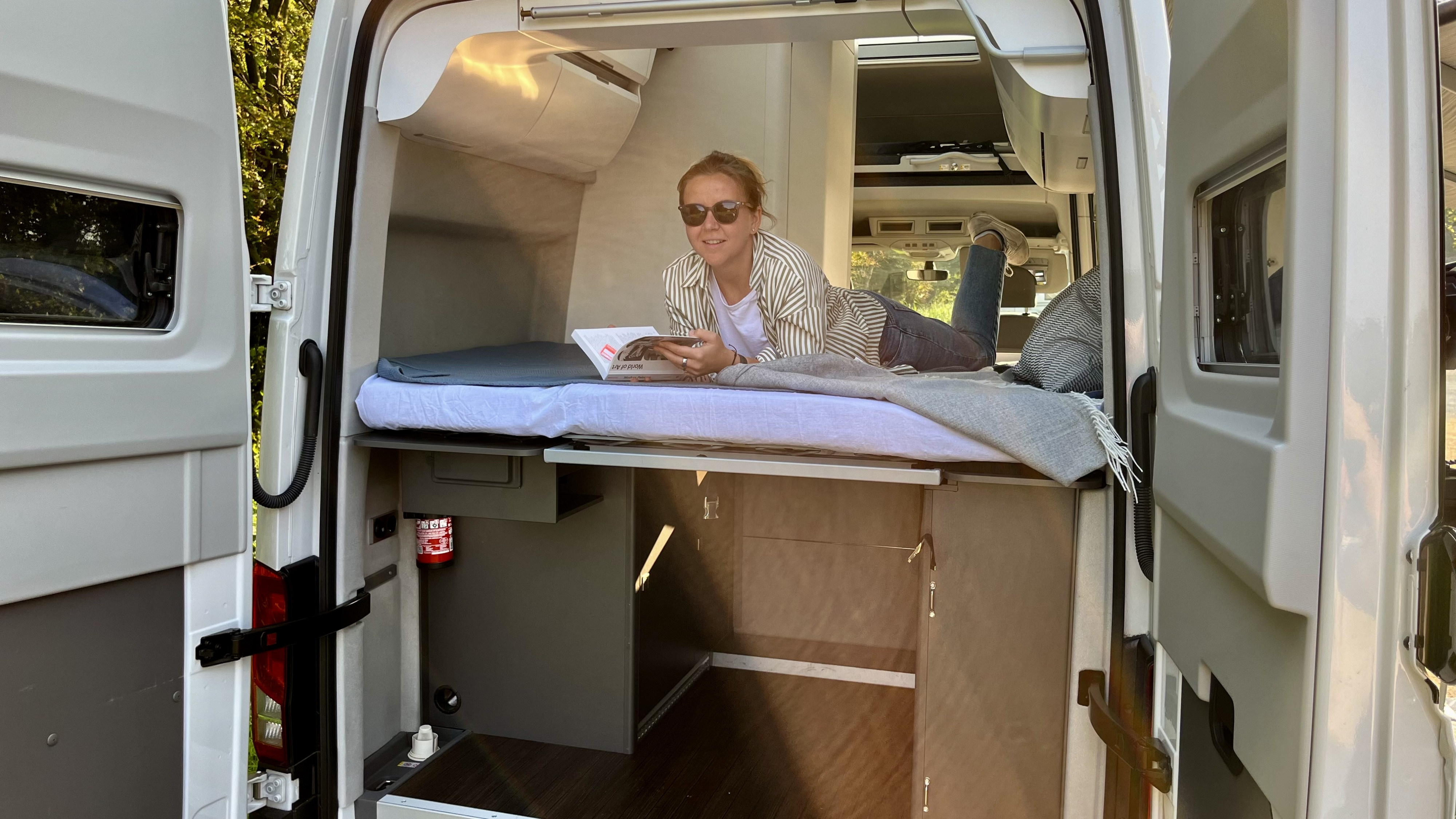 Woman relaxes reading in this RV rental camper van. Open rear doors showcase cozy bed, clever storage, and views.. Volkswagen Grand California - Clyde 2022
