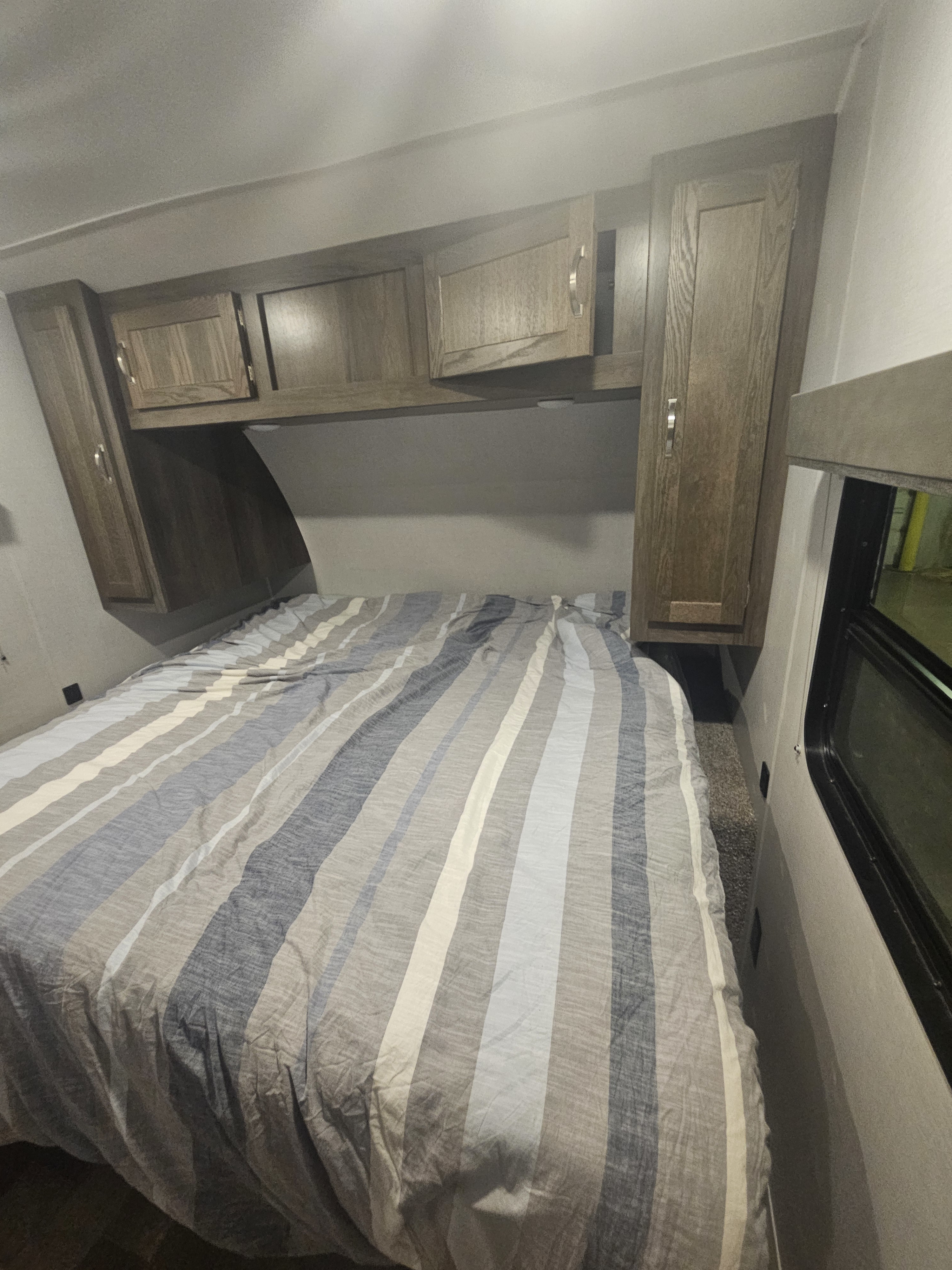 Compact RV rental bedroom featuring a striped bed, overhead wooden cabinets, and a narrow walkway by the window.. Forest River east to west 2020
