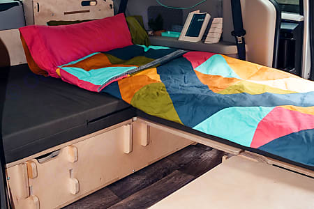 Cozy camper van bed with colorful bedding and integrated storage, perfect for an RV rental adventure. A compact and functional interior.. Chrysler Grand Caravan 2025