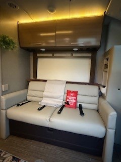 RV rental interior: A light-colored sofa with seatbelts, ready for travel. Folded wall bed and overhead cabinets.. Thor Gemini 2018