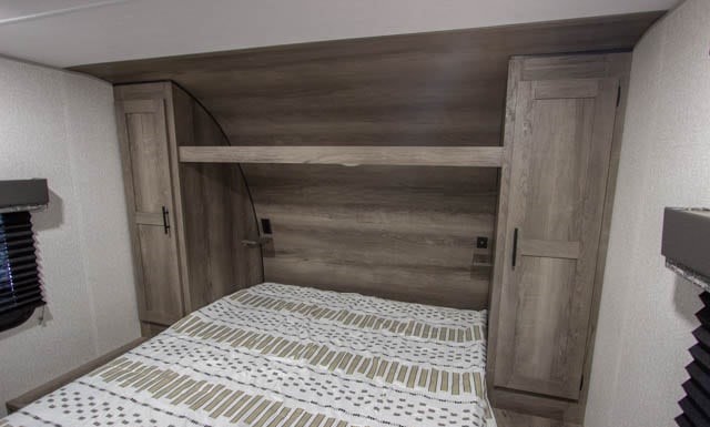 This RV rental's bedroom features a bed with striped bedding, an overhead storage shelf, and two wooden side cabinets.. K-Z Manufacturing Sportsmen 261BHKSE 2022
