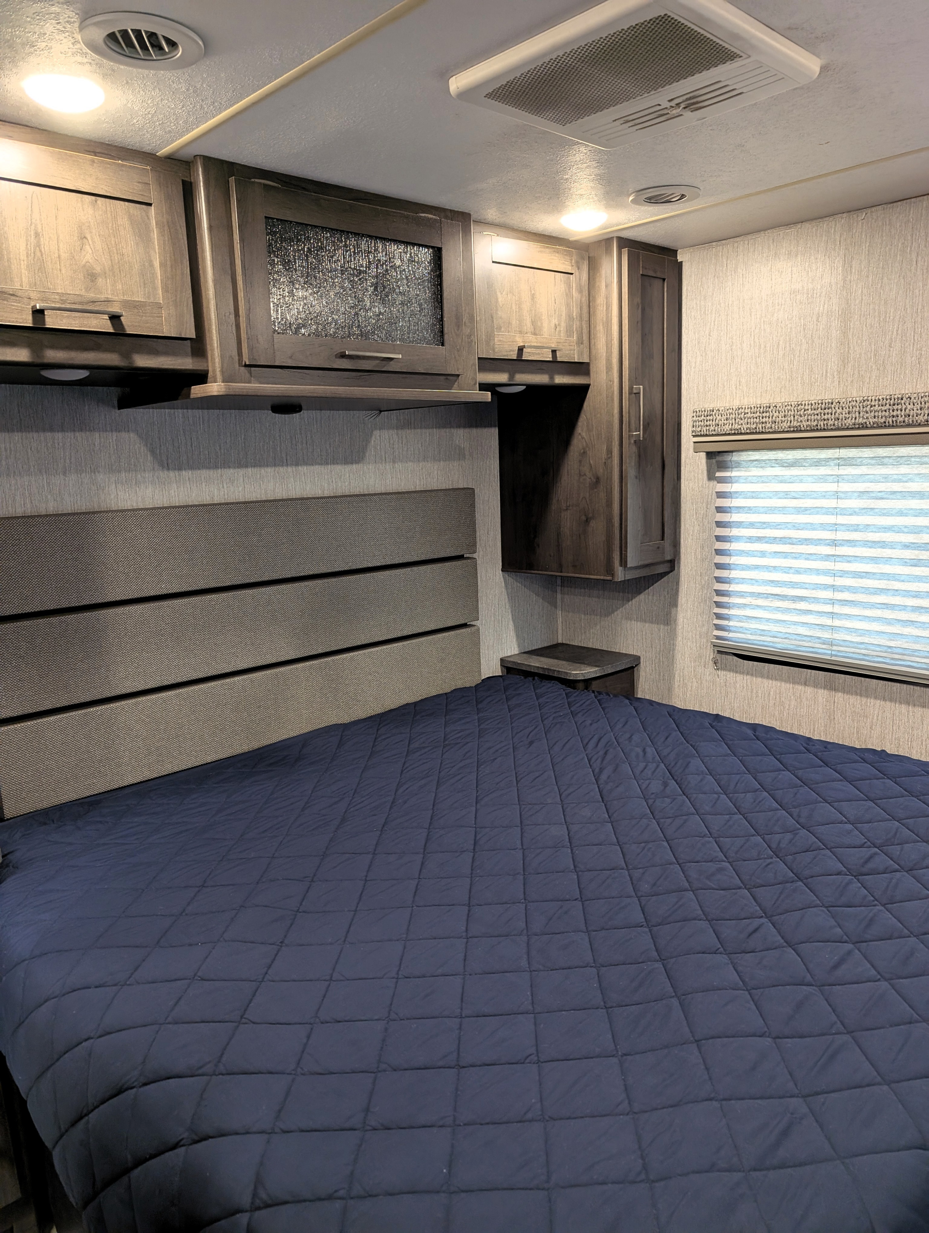 Comfortable RV rental bedroom featuring a dark blue bed, upholstered headboard, overhead cabinets, and AC unit.. Coachmen Pursuit 2022