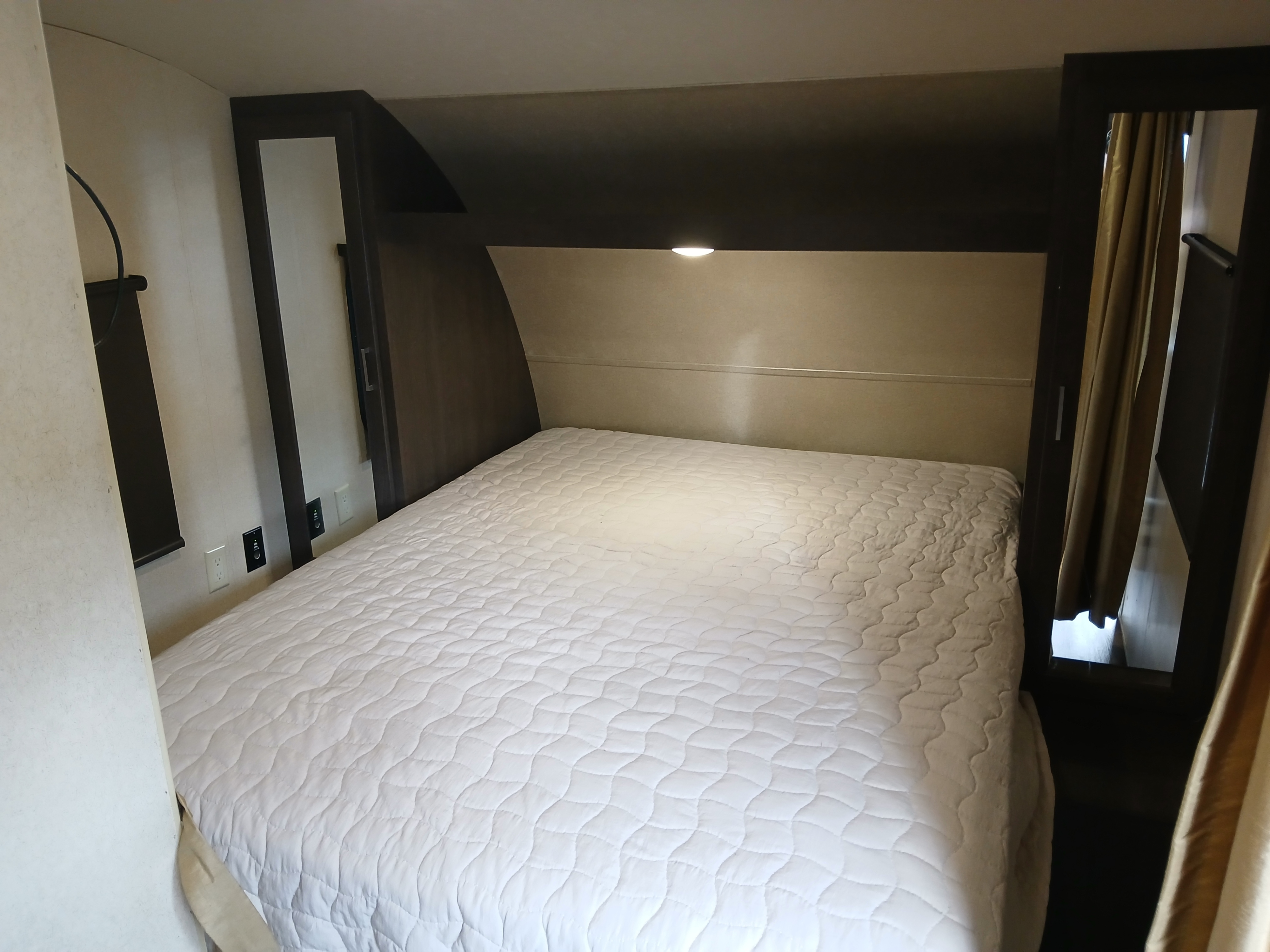 Inside an RV rental: a compact bedroom with a white quilted bed, built-in storage, and an overhead reading light.. Forest River Salem Cruise Lite 2018