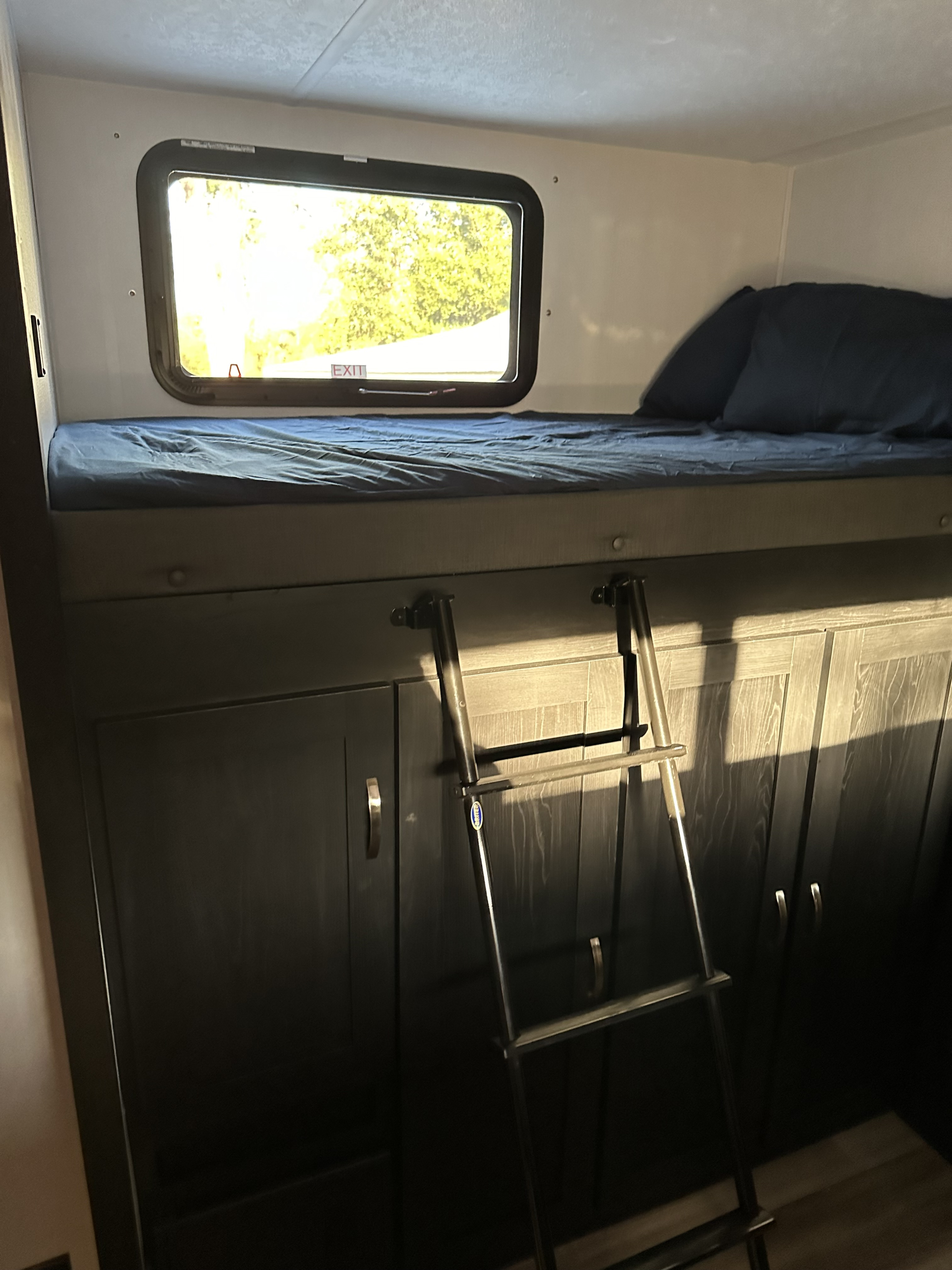 Inside an RV rental: a bunk bed with blue bedding, an exit window, a metal ladder, and dark wood-look storage cabinets below.. Coachmen 320BHDSLE 2023