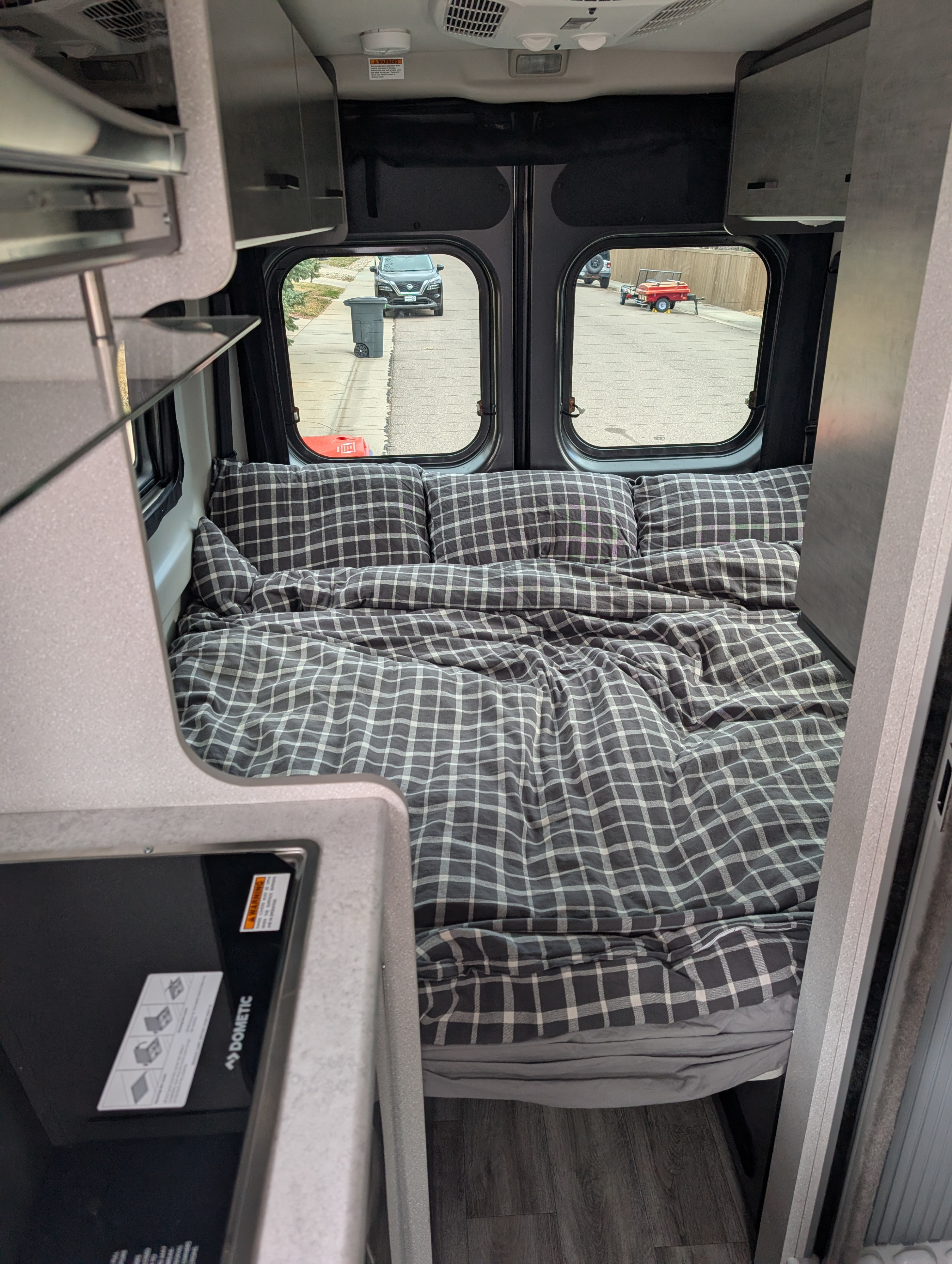 Inside a compact RV rental: a comfy bed with plaid bedding, overhead cabinets, and a street view through the rear windows.. Thor Dazzle 2024