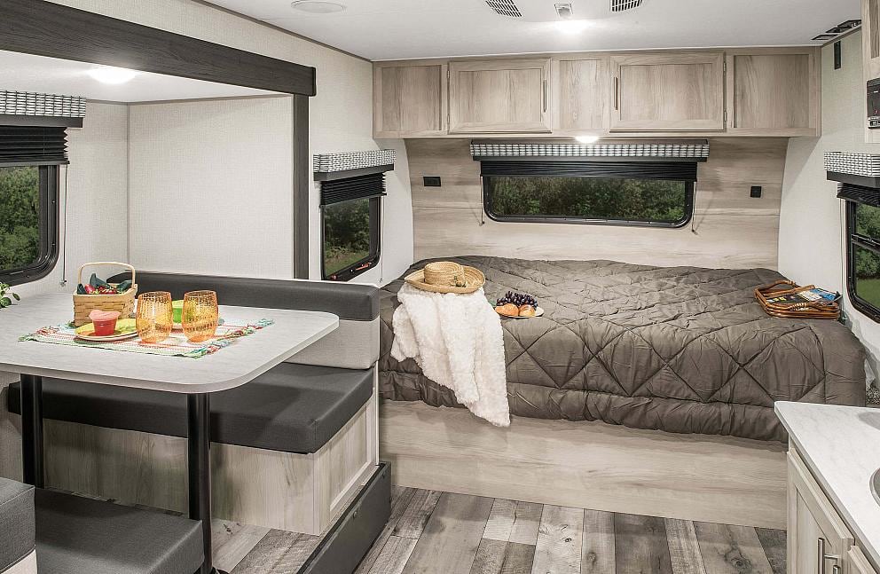 Inside a spacious rv rental, showing a well-lit dinette area with bench seating and a cozy bed with overhead cabinets.. KZ Escape 2021