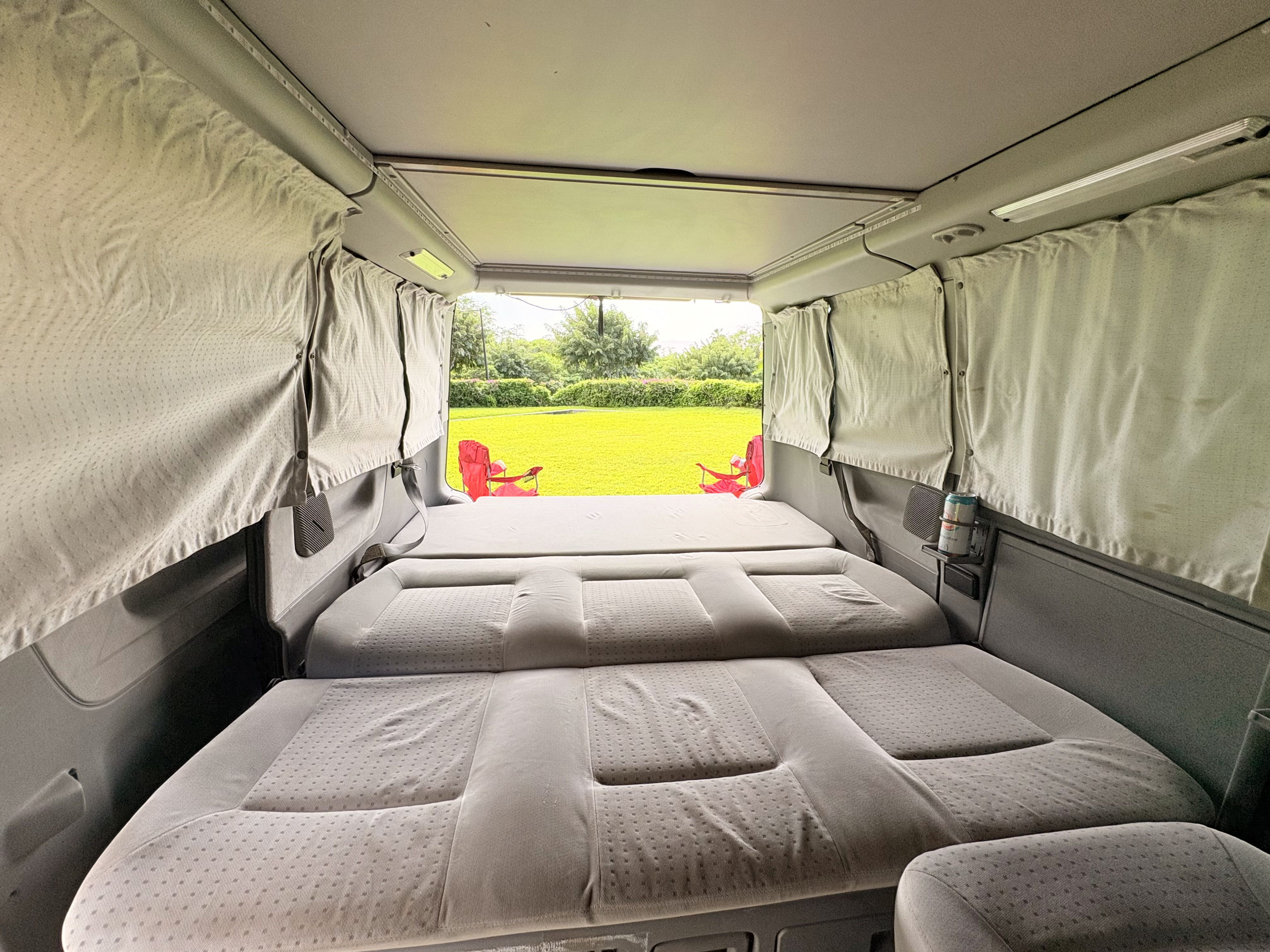 RV rental camper van interior. Rear seats fold into beds, offering a serene view of green nature and red chairs.. Volkswagen T4 Transporter 2002