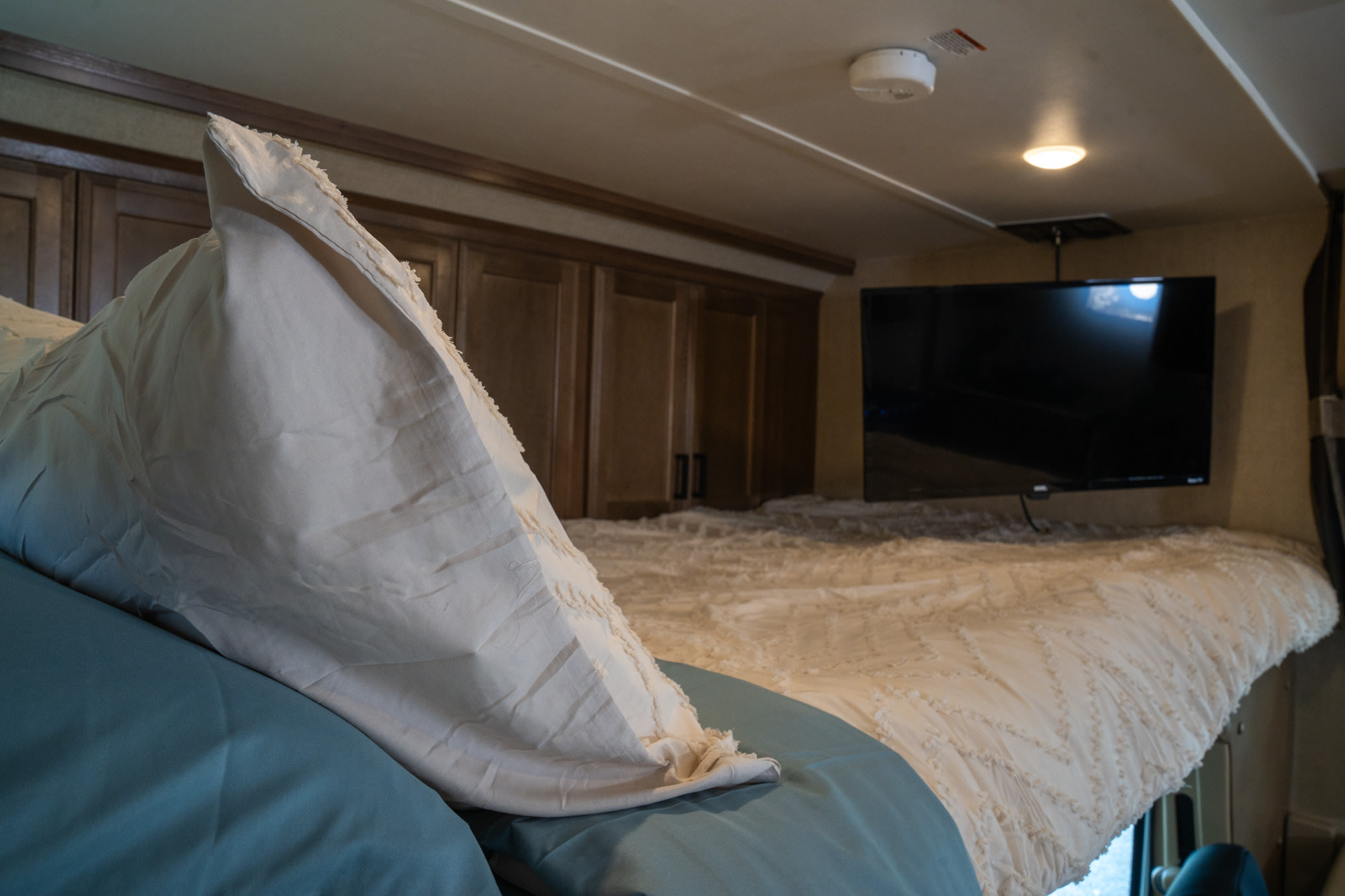 Comfortable cab-over bed in an RV rental, featuring a white comforter, blue sheets, mounted TV, and cabinets.. Gulf Stream Conquest 2019