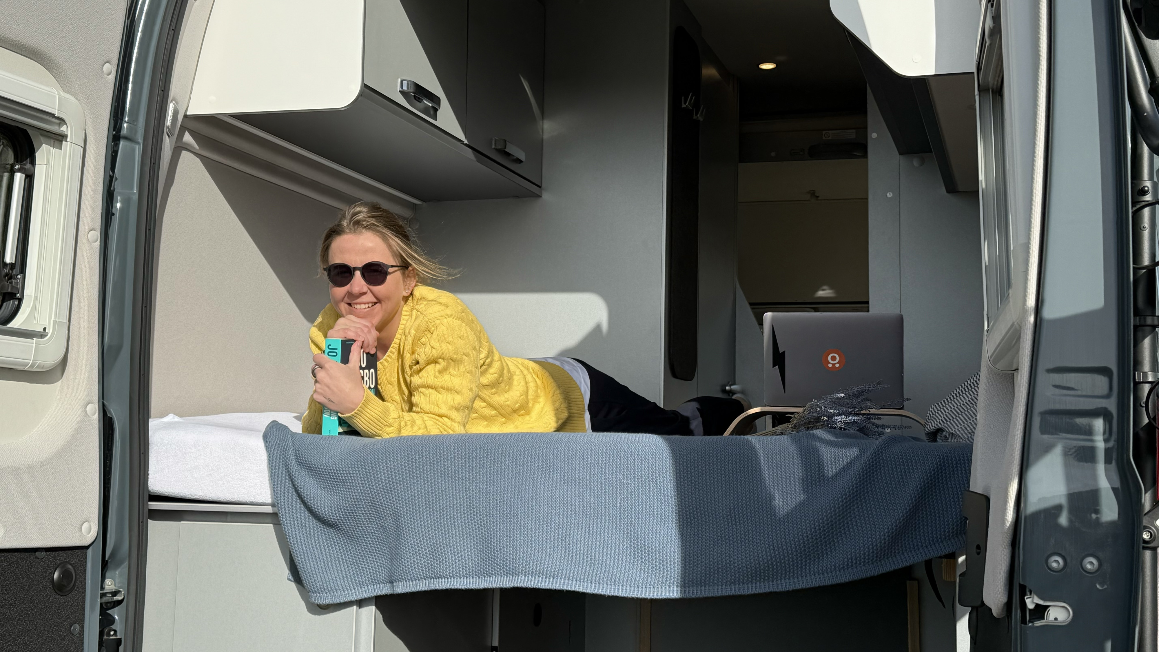 RV rental interior: Smiling woman in sunglasses relaxes on the bed, reading a book. Laptop visible.. Weinsberg DQ 2024