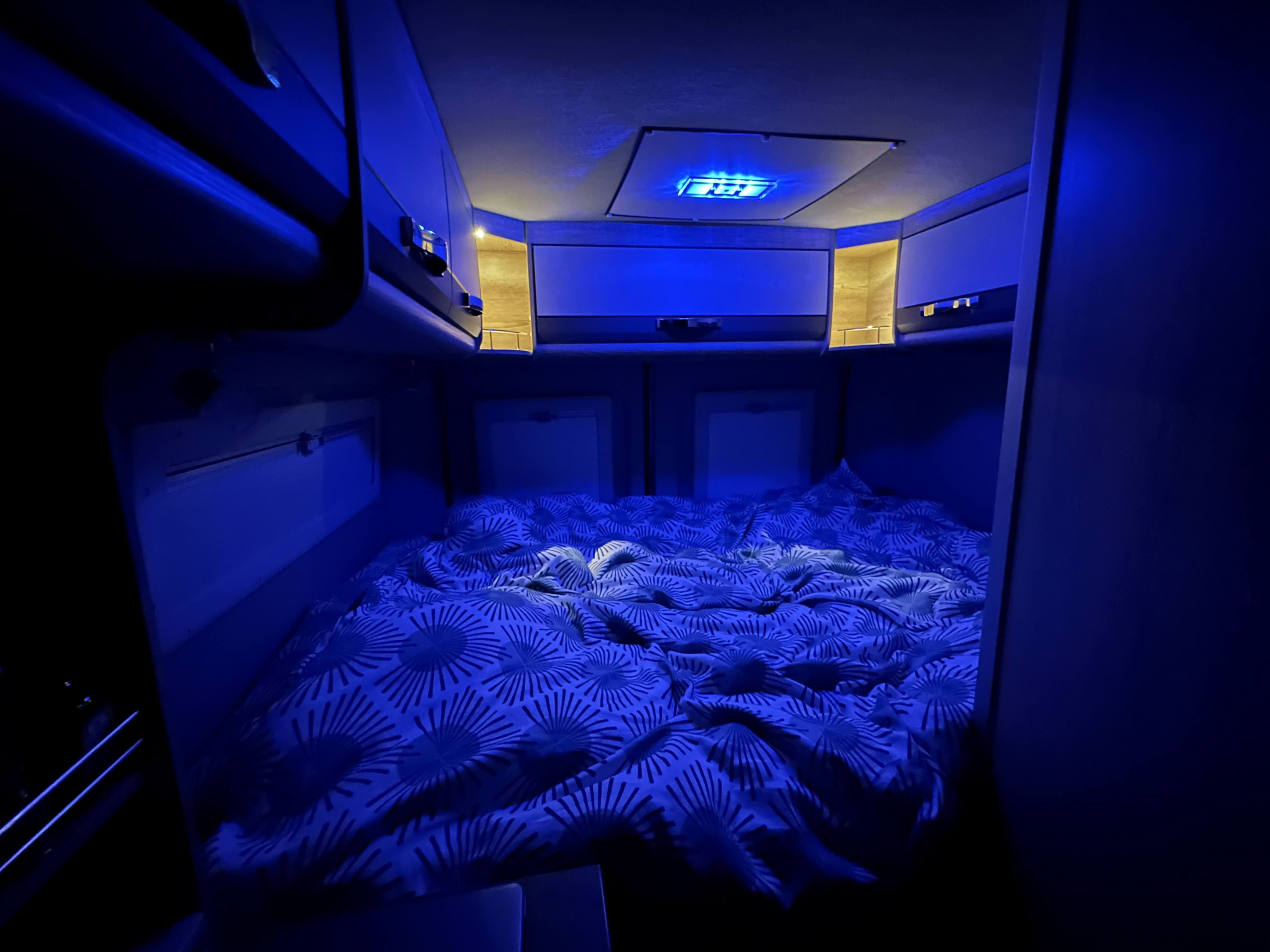 Cozy RV rental bedroom illuminated by ambient blue lighting, featuring a neatly made bed with a patterned duvet.. Pössl SUMMIT PLUS 2025