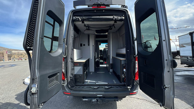Explore this RV rental's versatile camper van interior, featuring open rear doors, built-in storage, and a convertible bed.. Winnebago Revel 2025