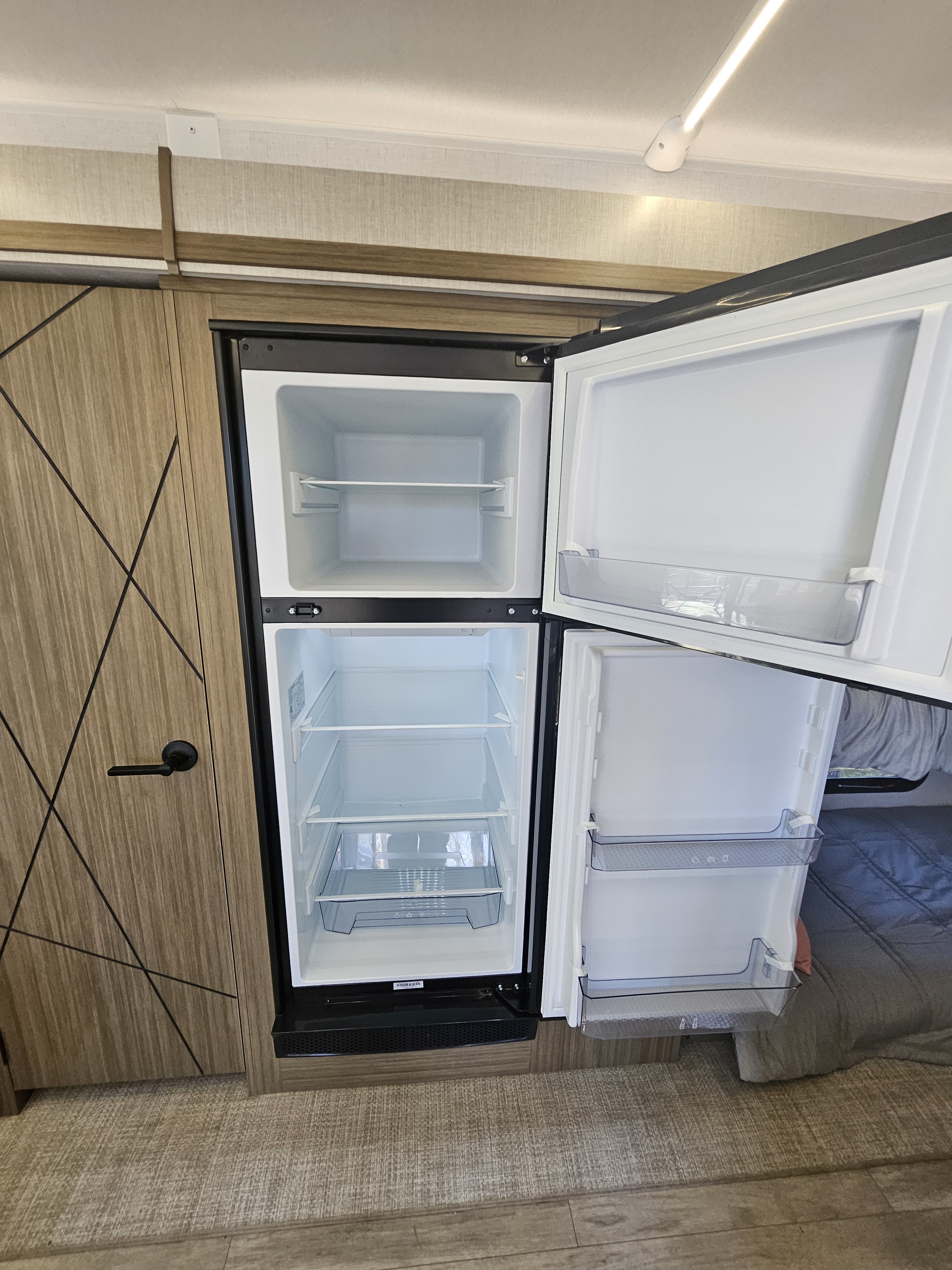 Inside an RV rental: a clean, empty two-door refrigerator/freezer with open doors, showing interior shelves, alongside cabinetry and a bed.. Forest River Wildwood 34Veranda 2025