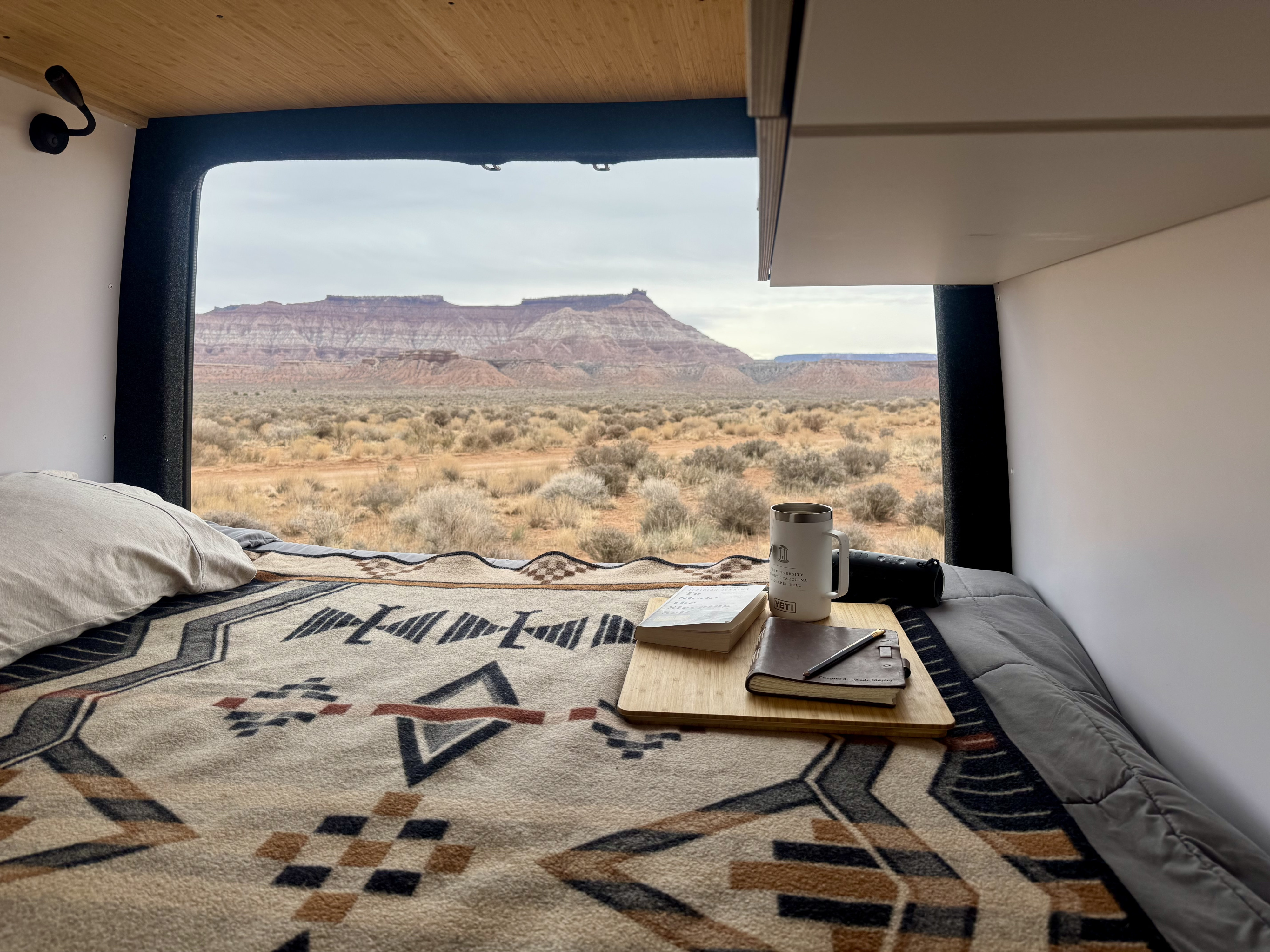 Cozy RV rental interior: Bed with patterned blanket, books, and coffee mug, overlooking a vast desert and mountains.. Mercedes Sprinter 2019