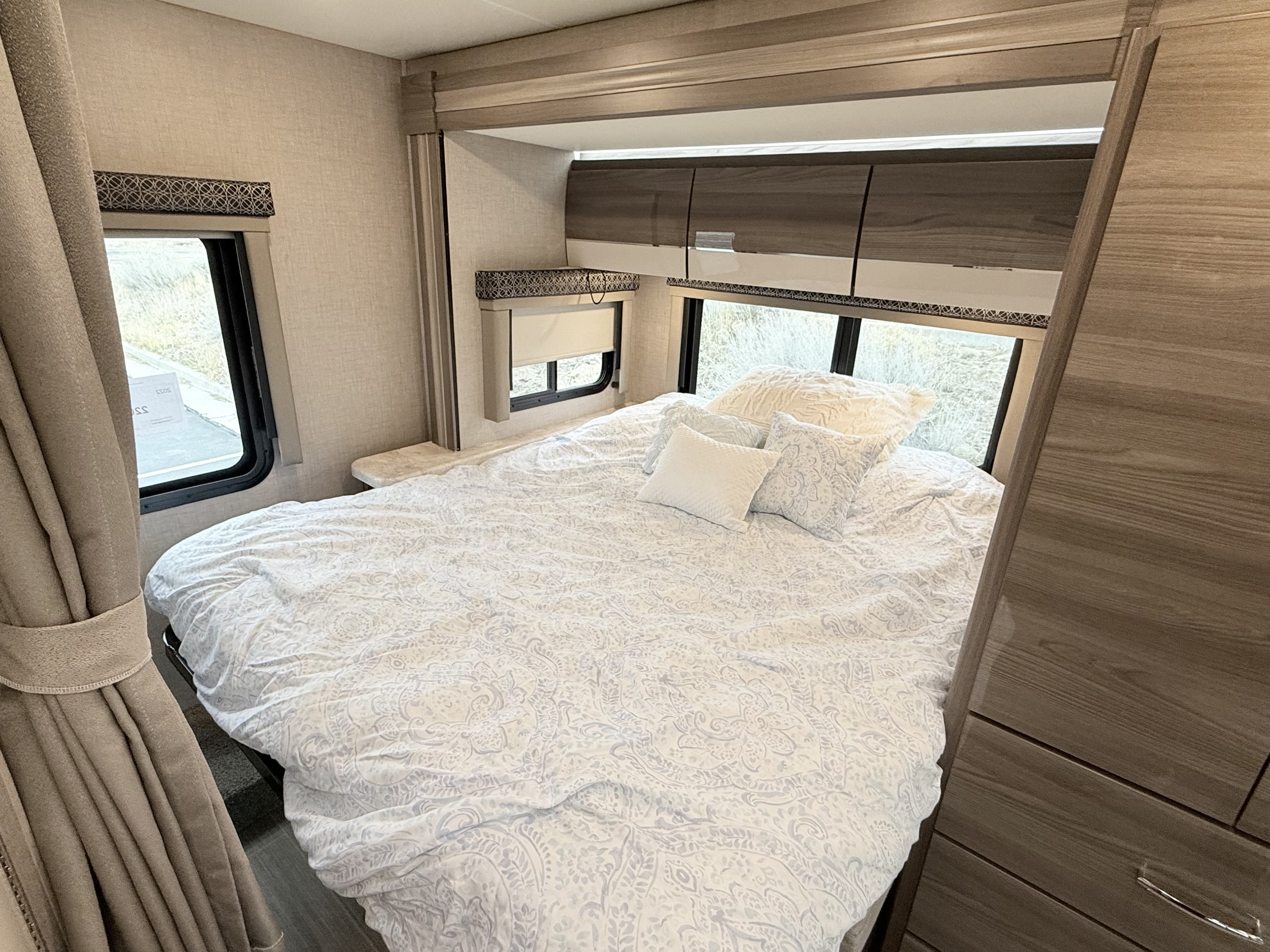 Cozy RV rental bedroom: a large bed with patterned white bedding, pillows, and bright windows with shades & curtains.. Thor Tiburon 2022
