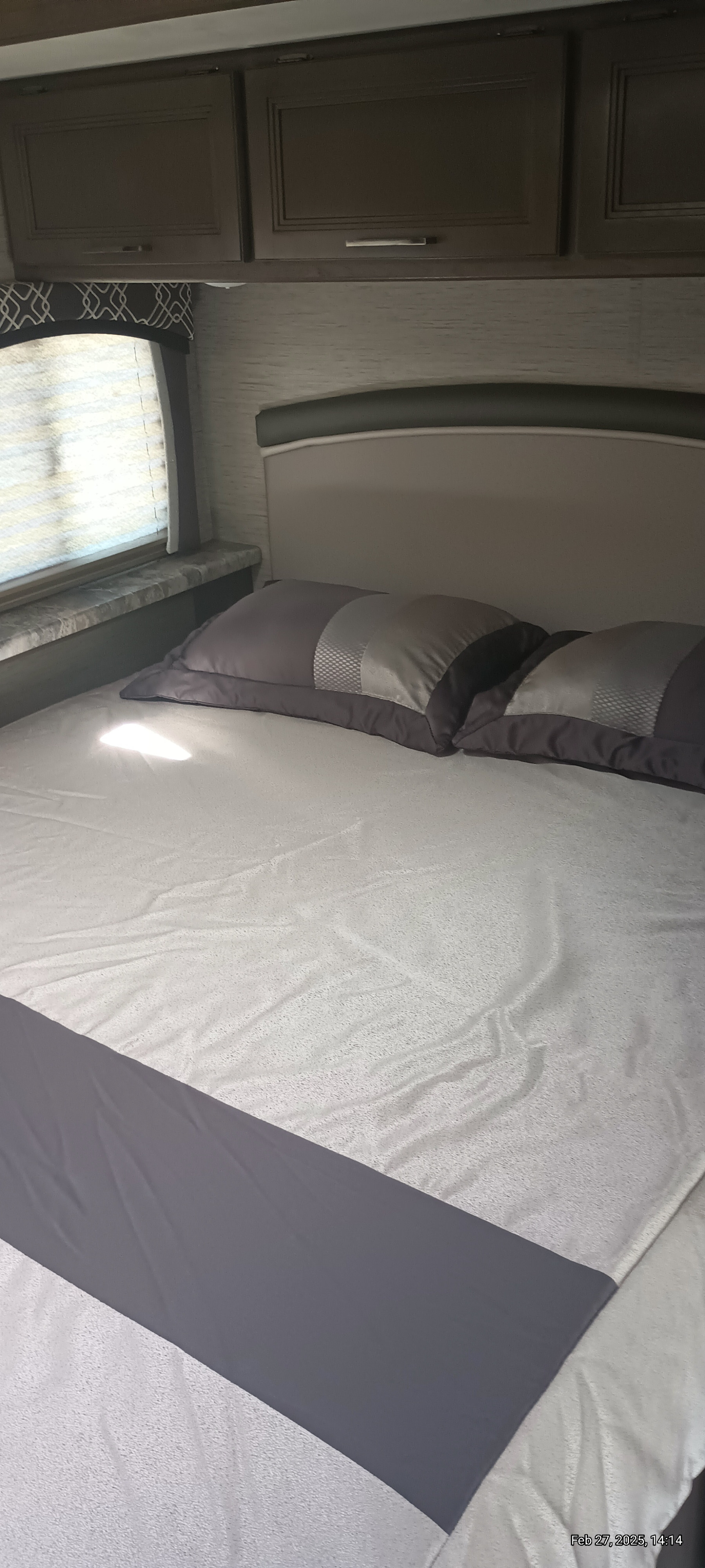 Cozy RV rental bedroom features a comfy bed with gray pillows, overhead cabinets, and a window for natural light.. Thor Motor Coach Chateau 2021