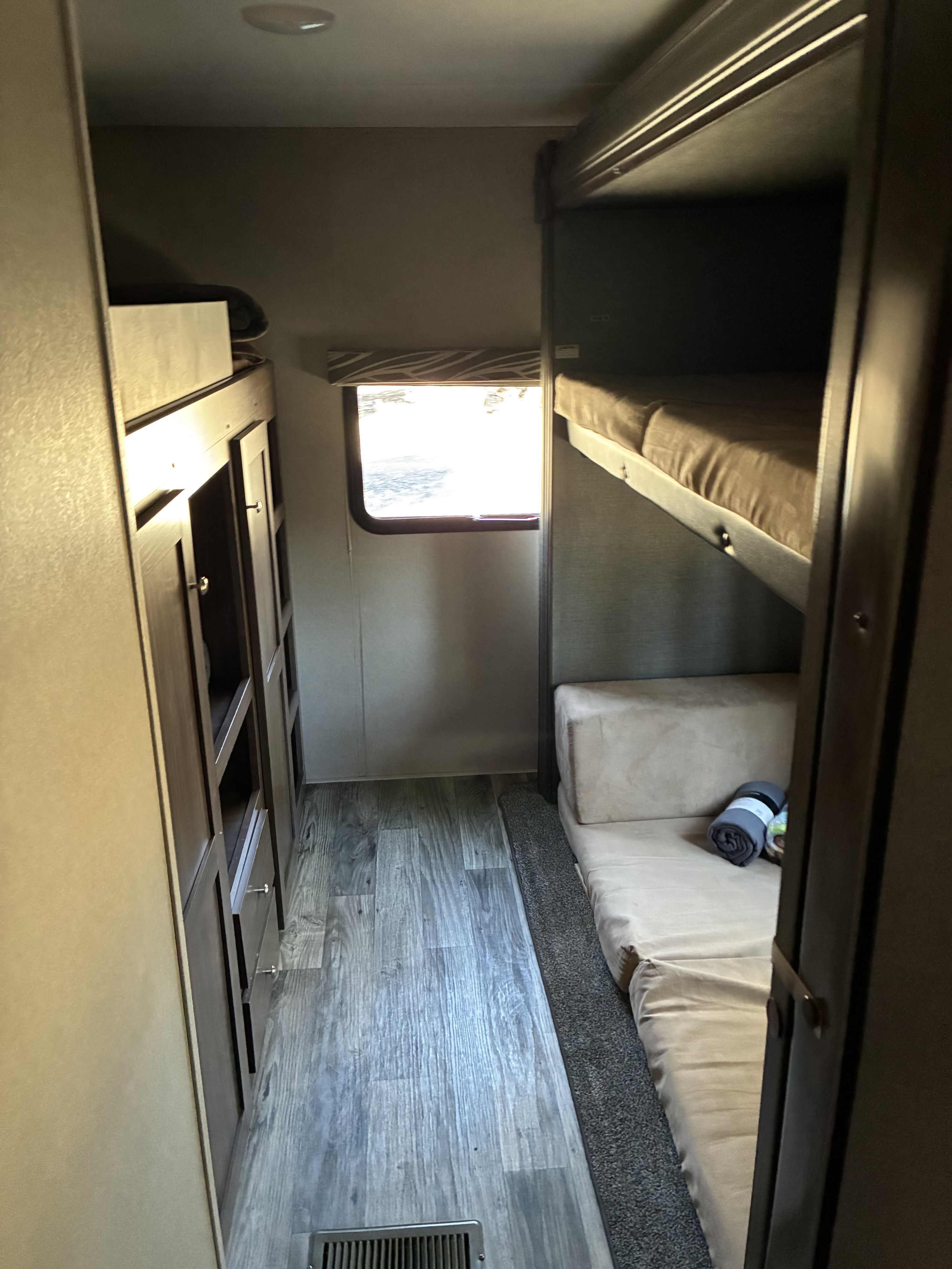 RV rental bunkhouse: a narrow corridor with dark wood bunk beds and storage on both sides, and a bright window at the end.. Keystone Hideout 2019