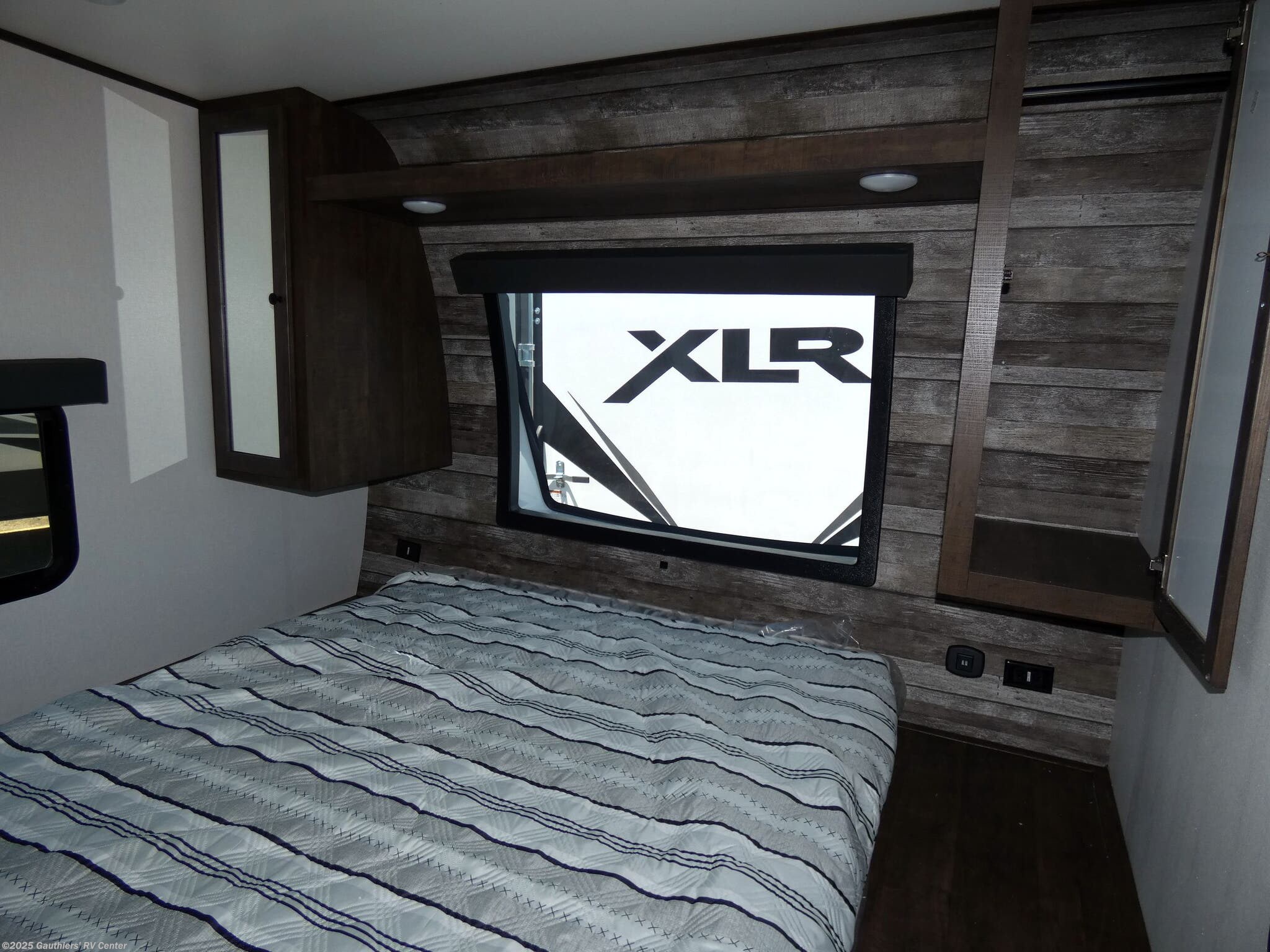 RV rental interior: a compact bedroom with a cozy striped bed, window displaying 'XLR' logo, and built-in storage.. Forest River XLR Hyperlite Toy Hauler 2023