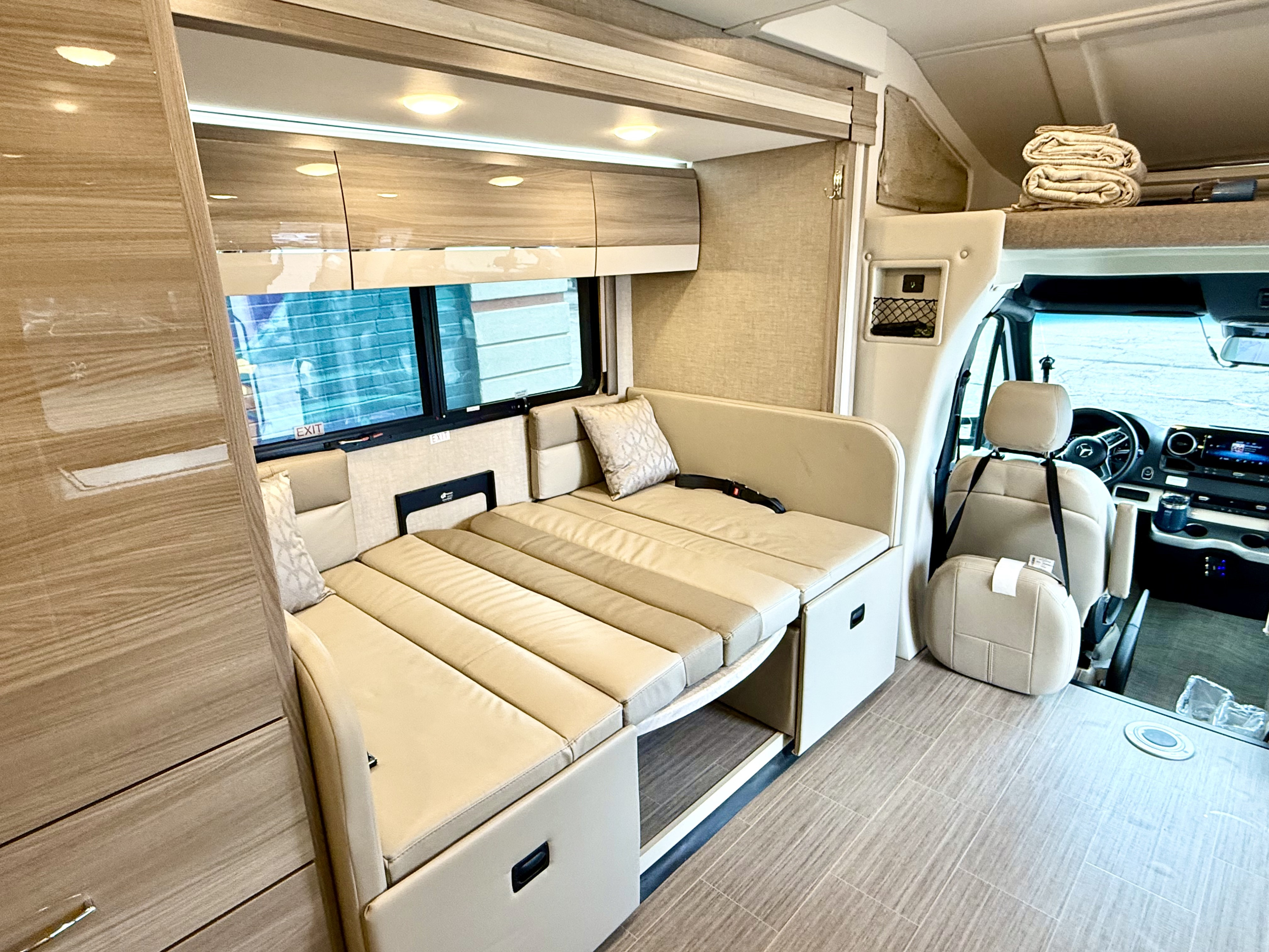 Modern RV rental interior featuring a beige convertible couch/bed, wood cabinets, and driver's cab.. Thor Delano 2023
