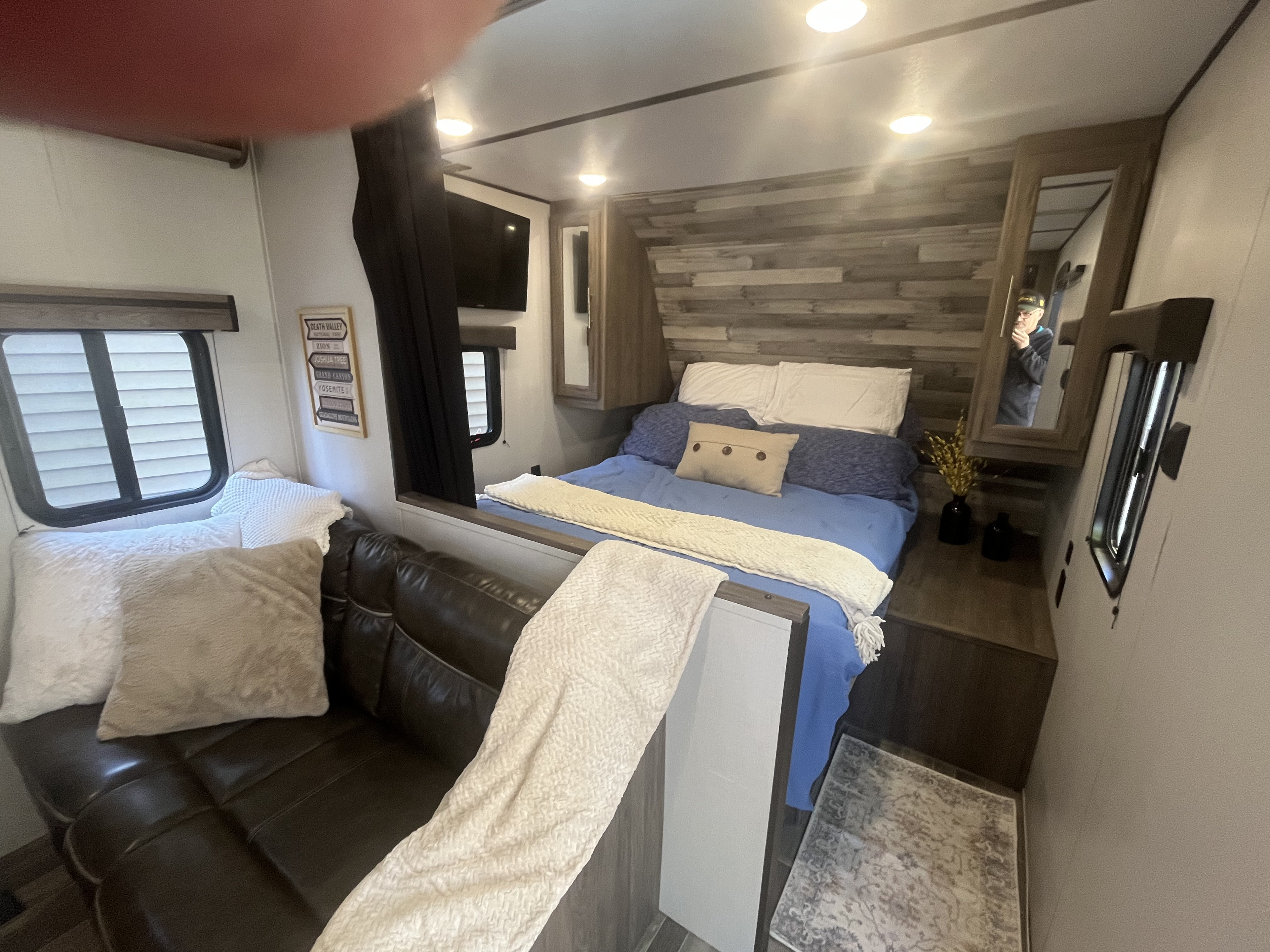 Cozy rv rental interior. Features a bed with blue bedding, a comfy leather sofa, and national park sign decor. Person reflected in mirror.. Crossroads RV Zinger Lite 2021