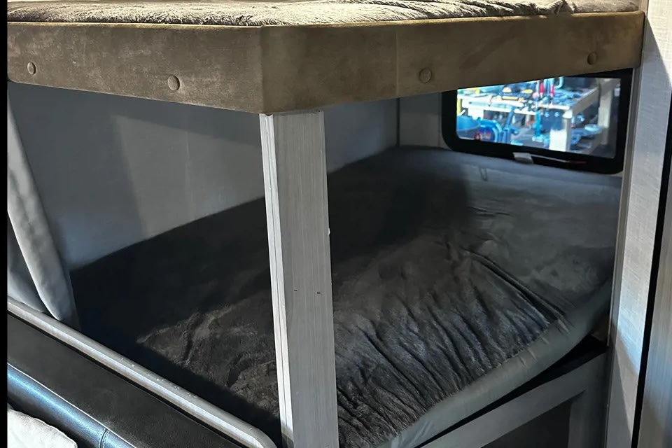 This RV rental interior features two bunk beds with soft grey bedding. A small window next to the lower bunk shows an outside view.. Keystone RV CM18BH22 2022