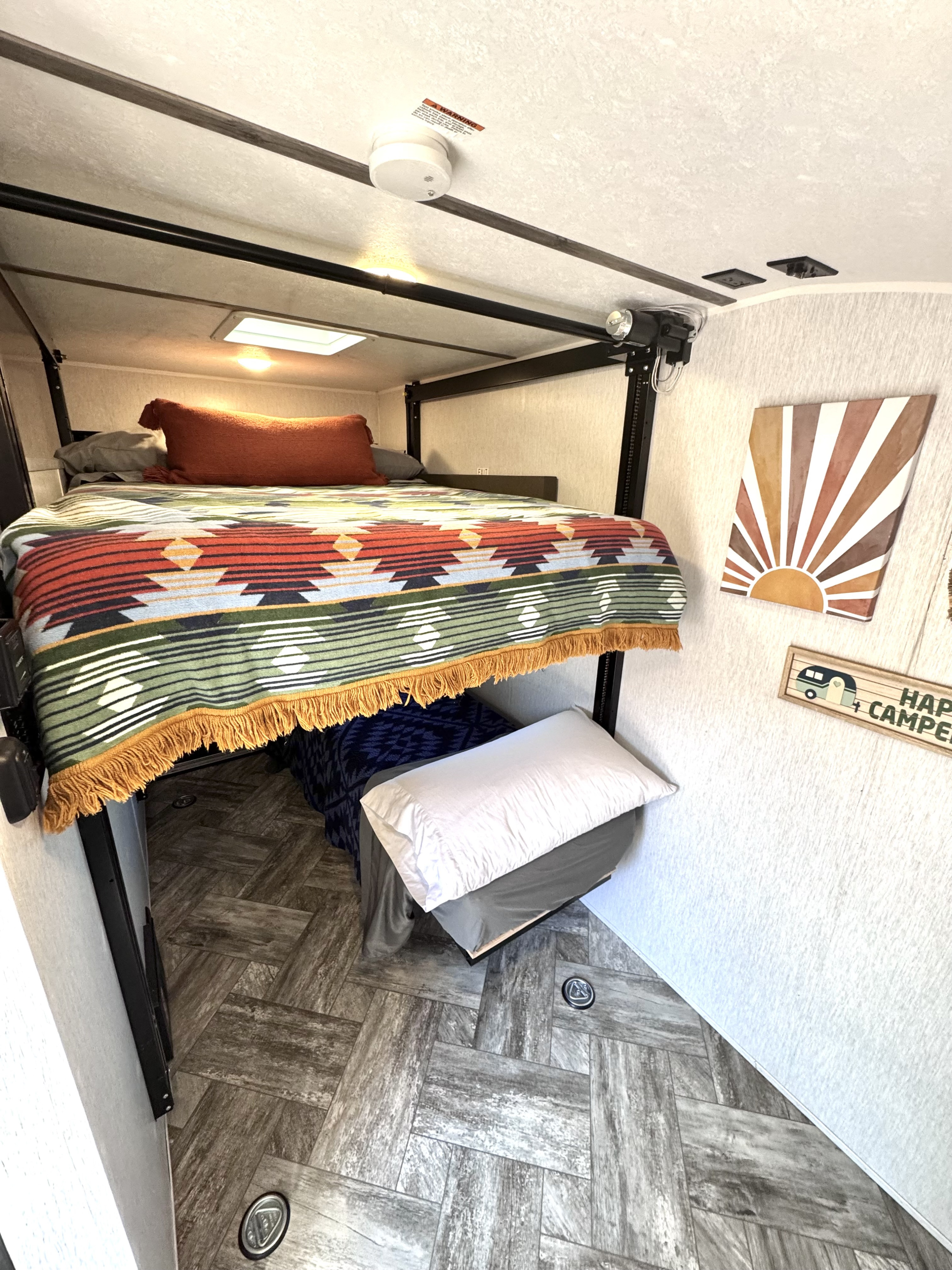 Cozy RV rental interior featuring an elevated bed with a vibrant blanket, wood-look floor, and charming camper decor.. Sunset Park RV SunRay 2024