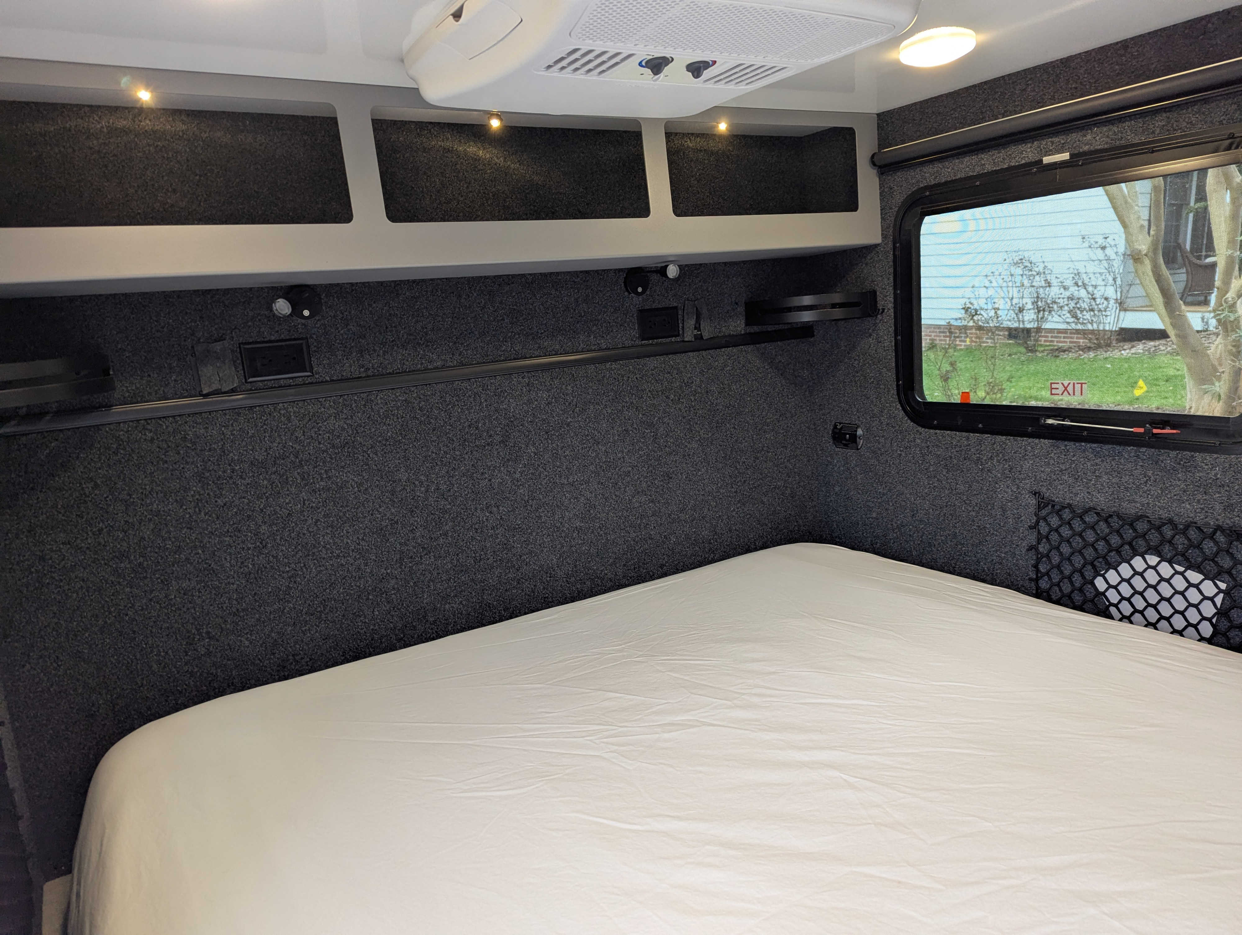 Cozy RV rental bedroom with a white bed, dark carpeted walls, illuminated overhead storage, and a window showing 'EXIT'.. Encore RV ROG 2022