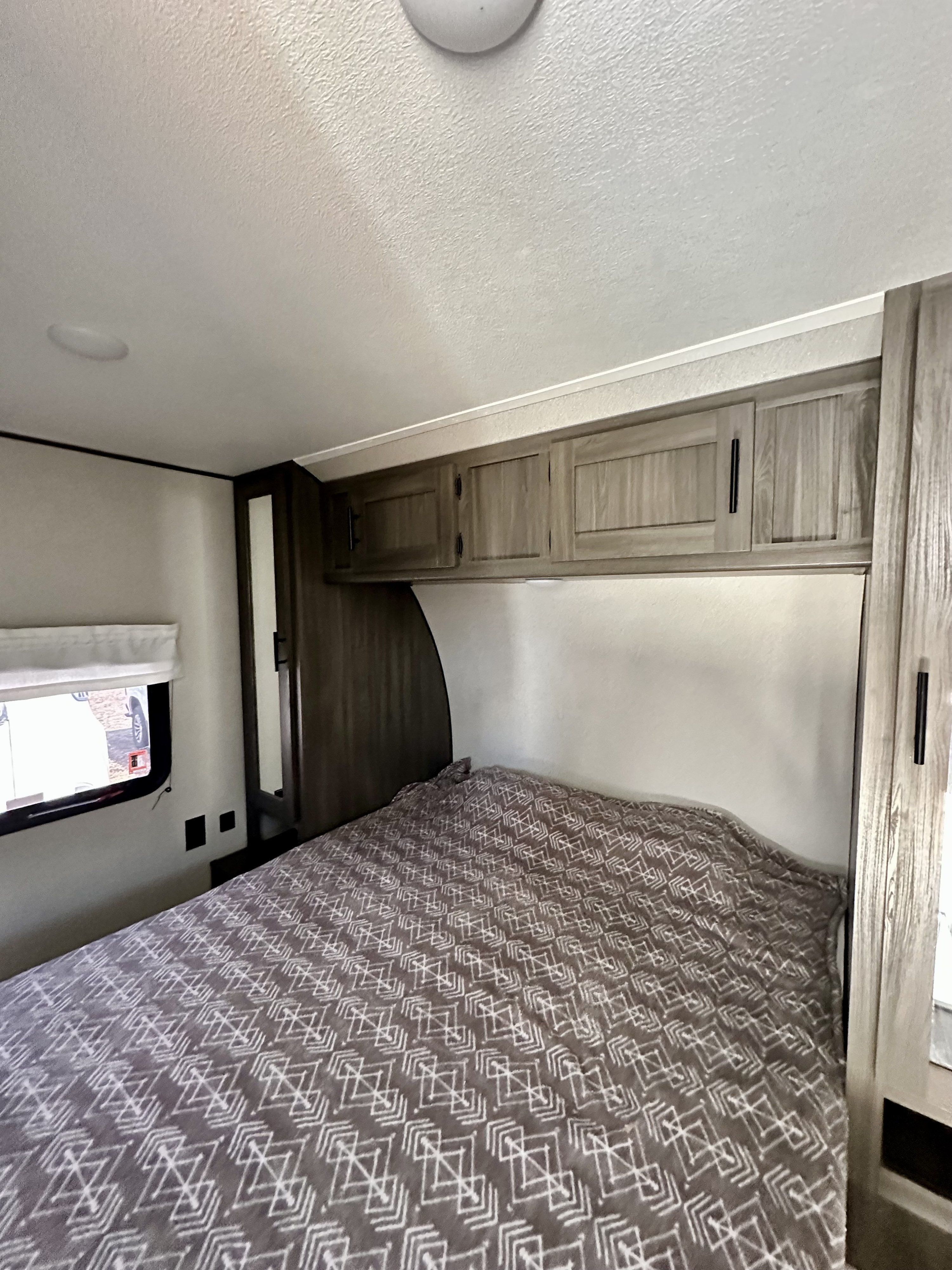 This RV rental bedroom features a cozy bed with patterned blanket, overhead cabinets, and a small window, ready for your next trip.. Forest River Shasta 2021