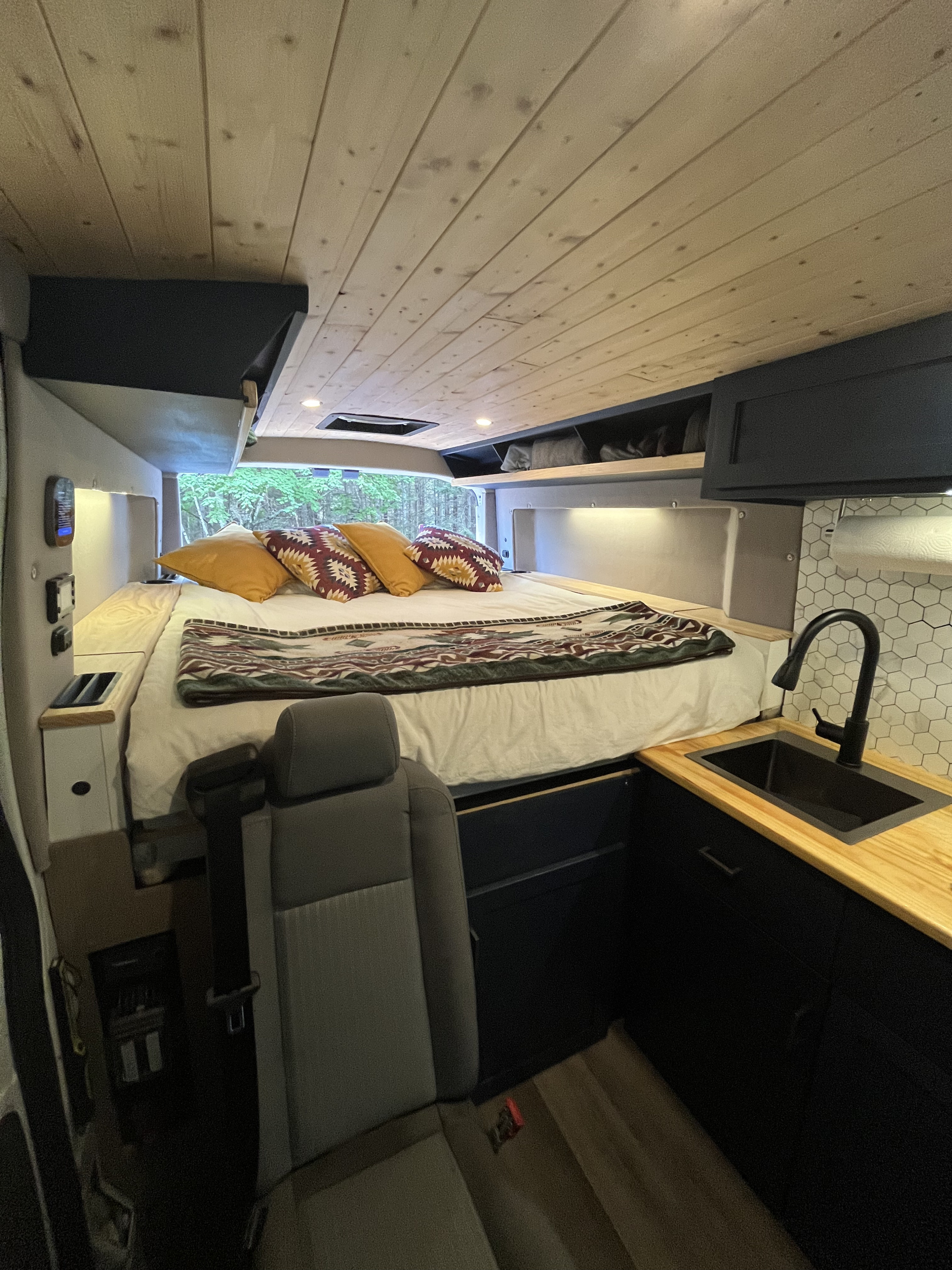 Cozy RV rental camper van interior. Features a raised bed looking into trees, wood ceiling, and compact kitchen.. Ford Transit Custom 2017