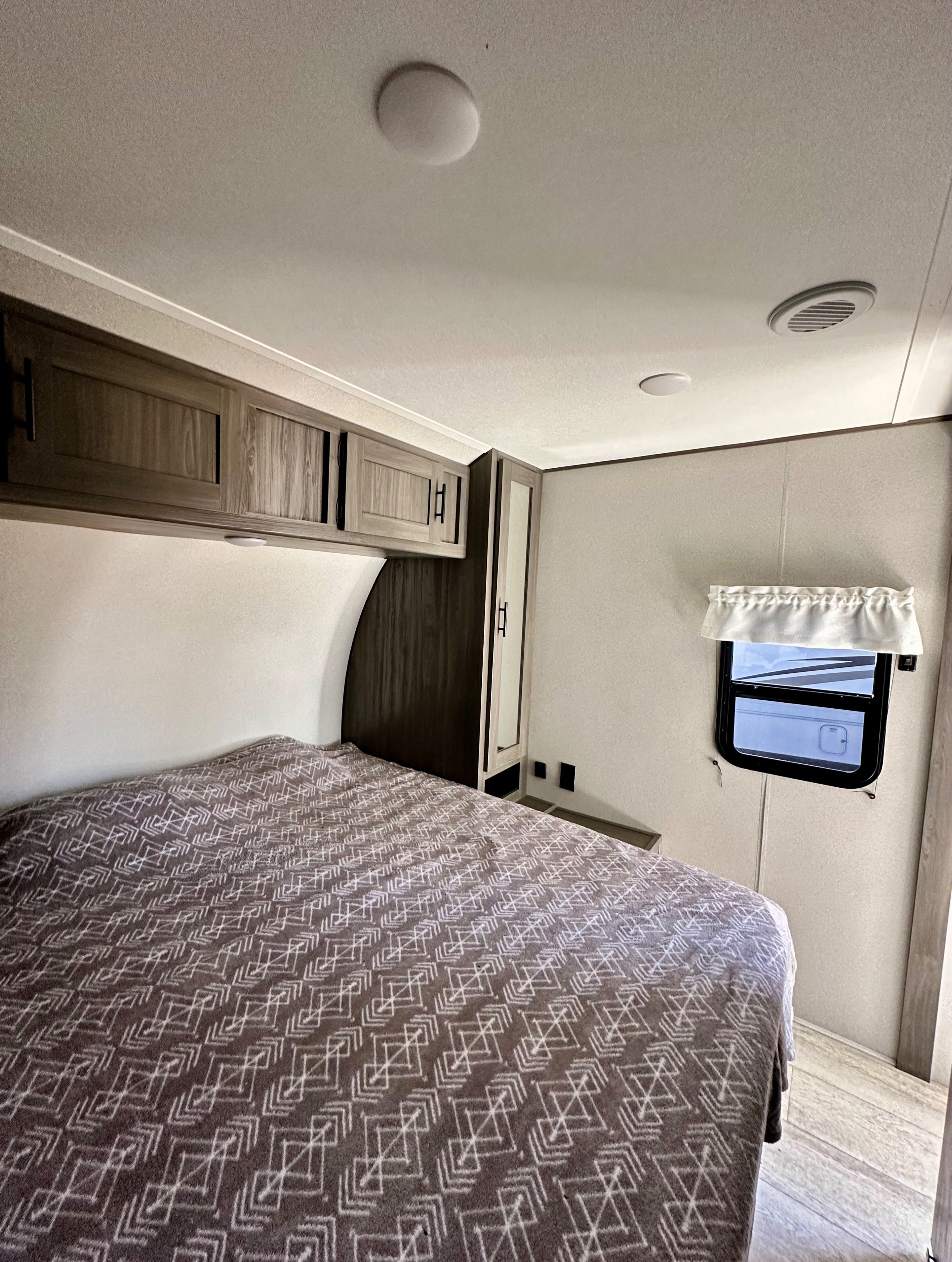 This RV rental bedroom offers a comfortable bed with a patterned blanket, overhead cabinets, and a side window with a valance.. Forest River Shasta 2021