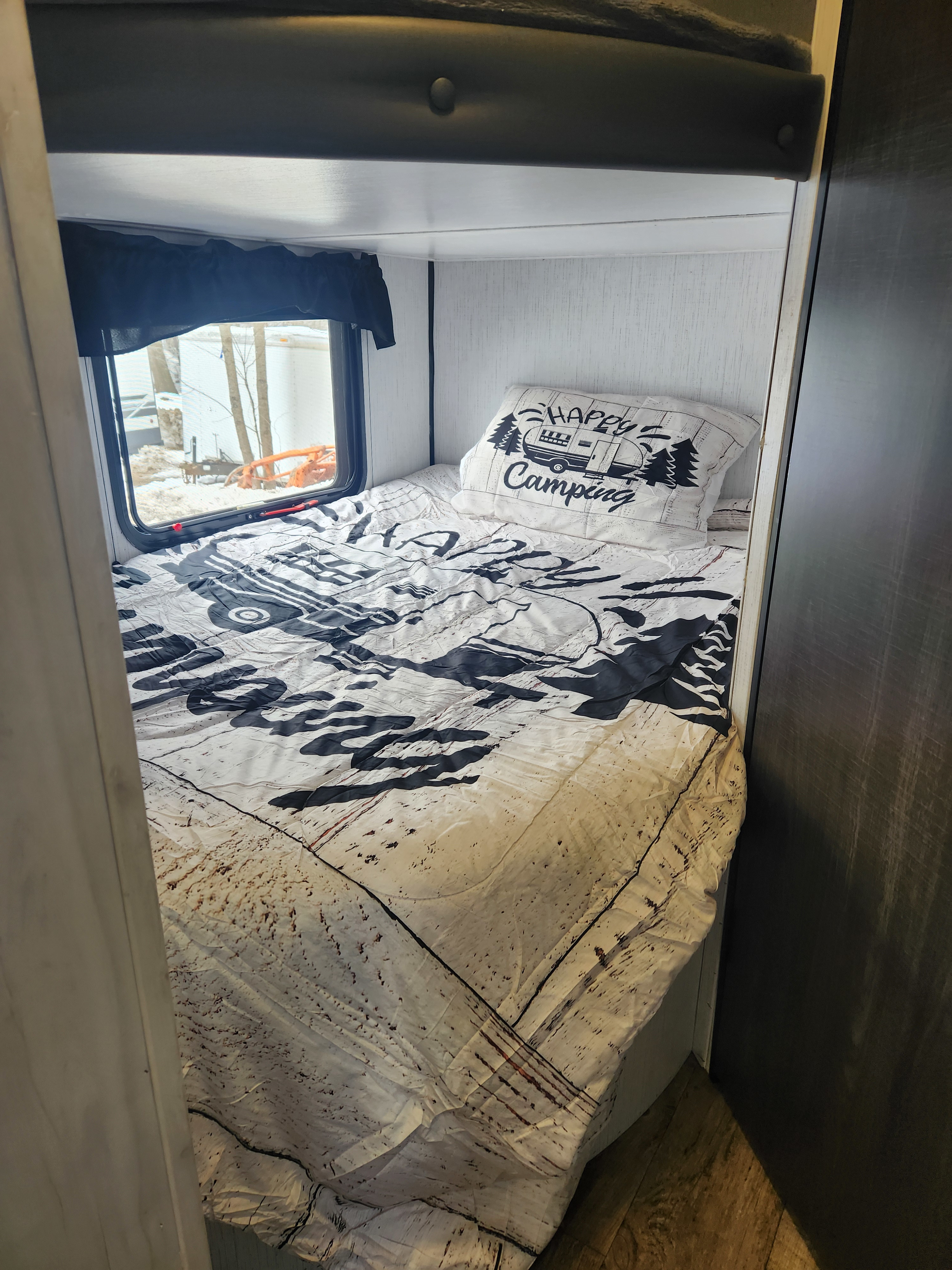 Cozy RV rental bunk bed with a 'Happy Camping' pillow, patterned bedding, and a window offering a snowy outdoor view.. Heartland RVs Trail Runner 2022