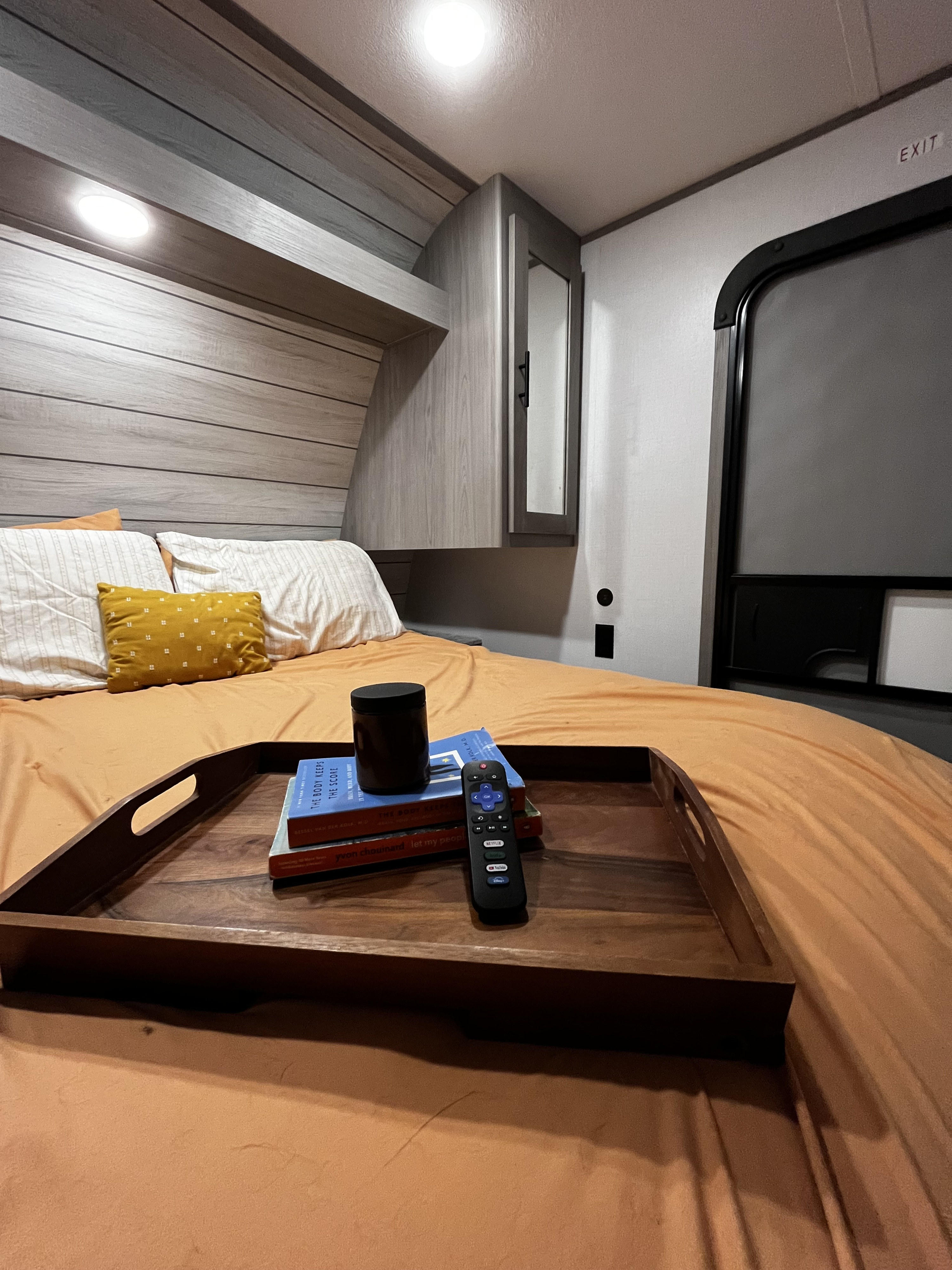 Cozy RV rental bedroom showing a bed, wooden tray with books & remote, and an exit door, offering a comfortable stay.. Dutchmen Coleman Lantern LT 2024