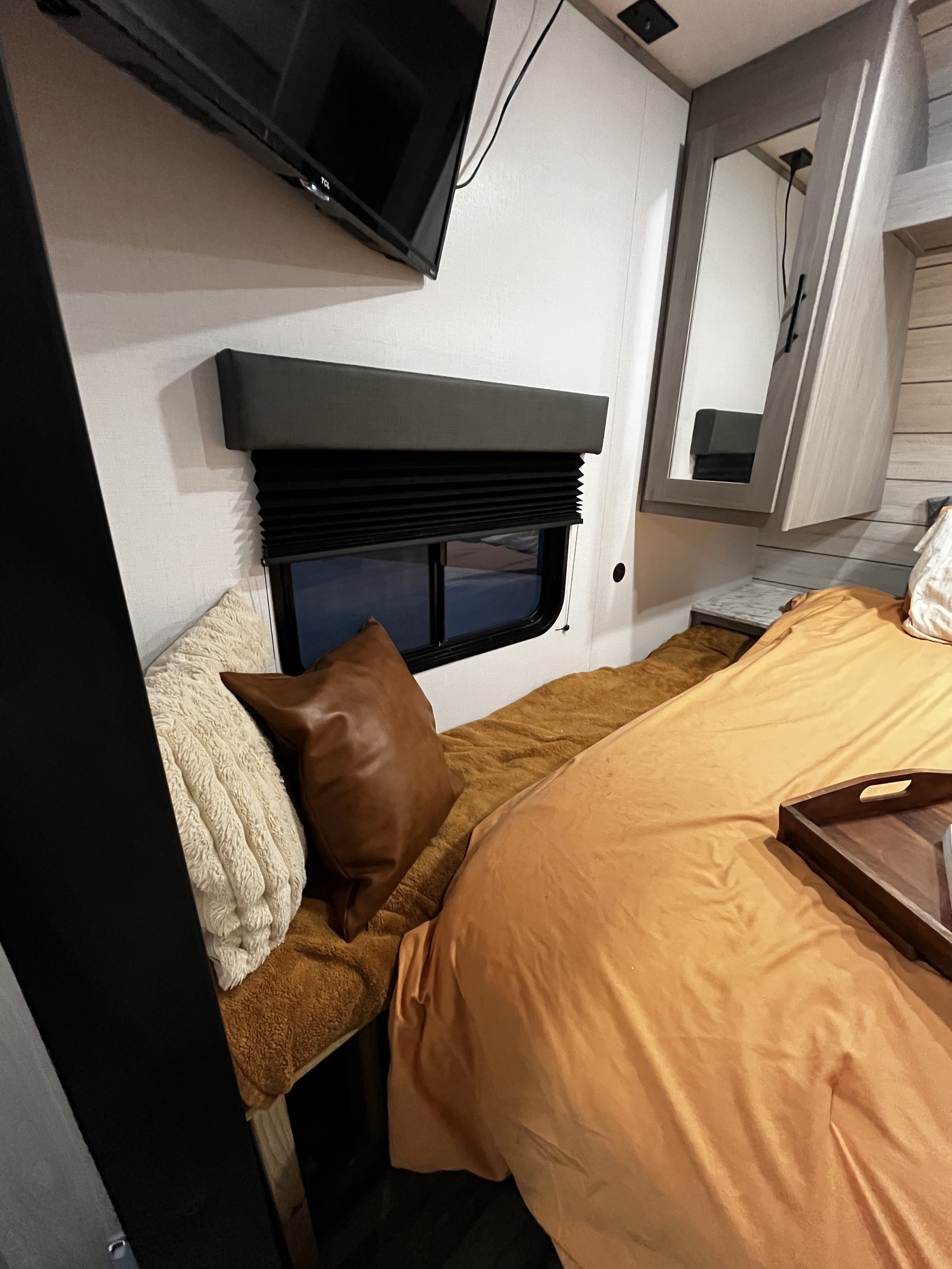 View inside an RV rental bedroom showing a cozy bed with orange sheets, a window with blinds, and a wall-mounted TV.. Dutchmen Coleman Lantern LT 2024