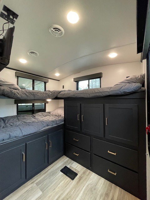 Inside an RV rental: two bunk beds with gray bedding & custom black storage cabinets. Bright interior lights & wood-look floor.. Keystone RV Outback Ultra-Lite 2022