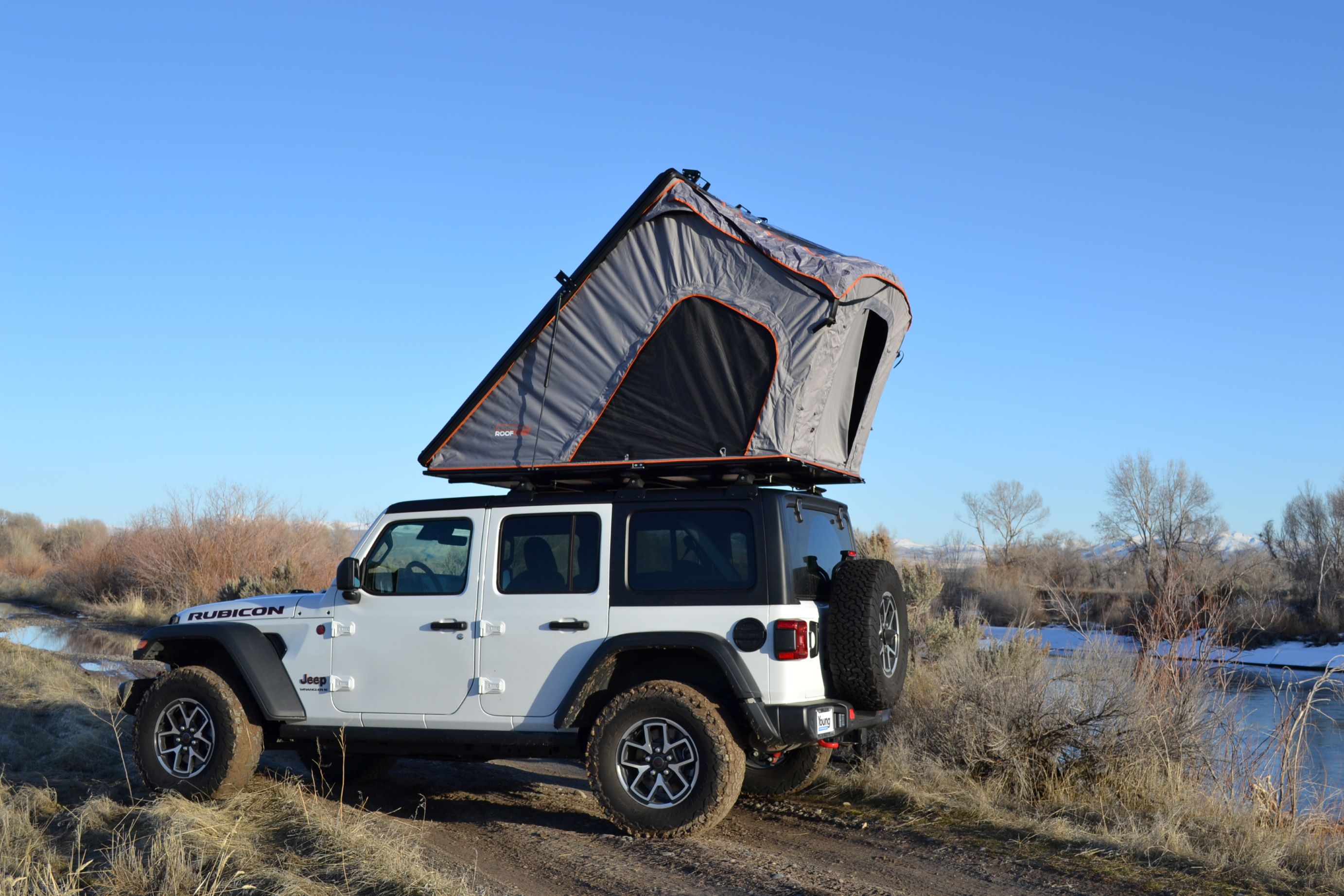 White Jeep Rubicon with grey rooftop tent deployed by a scenic winter river, perfect for an adventurous RV rental.. Jeep Wrangler Rubicon 2024