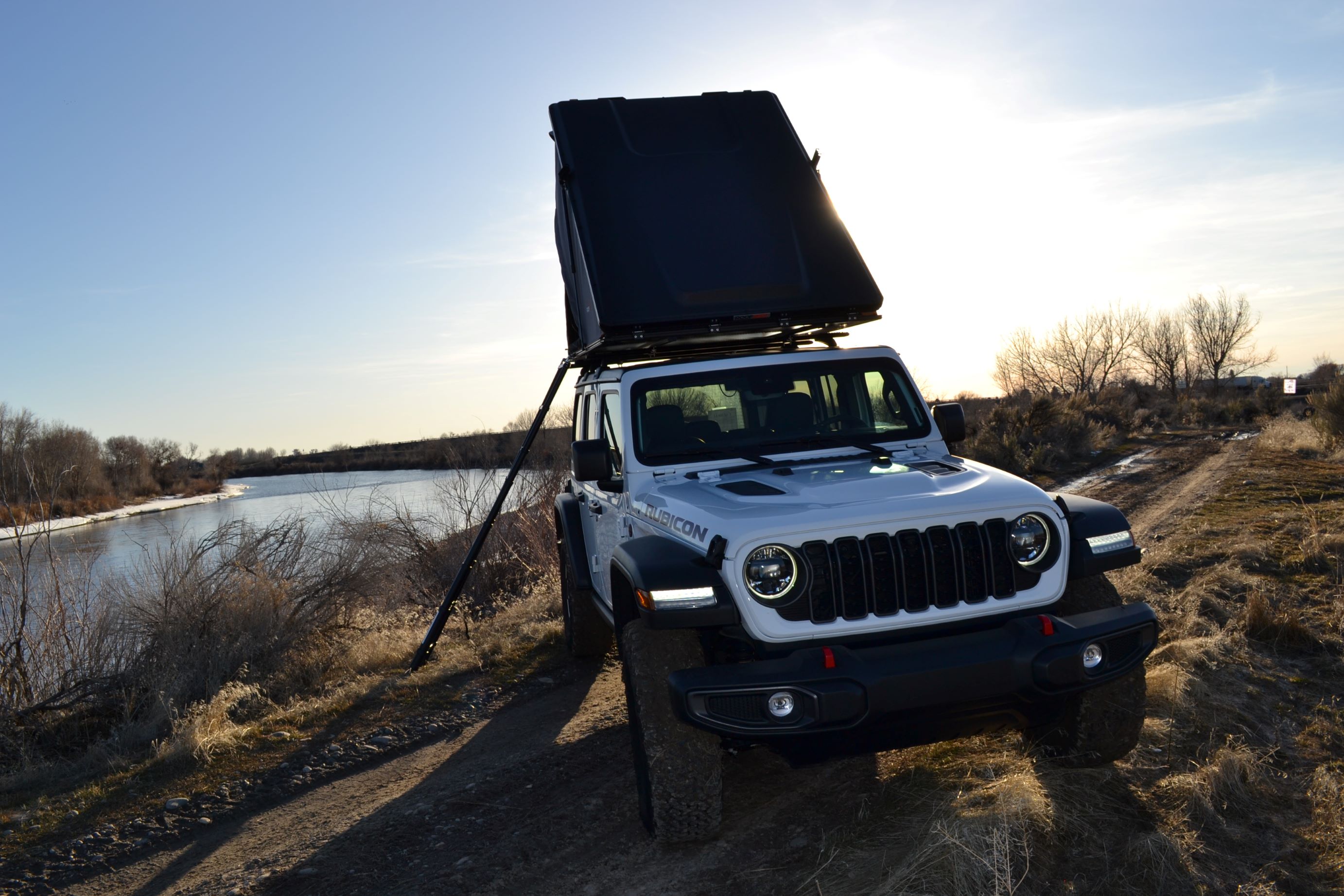 White Jeep Rubicon with a deployed rooftop tent, parked by a river at sunset. Ideal for an adventurous rv rental experience.. Jeep Wrangler Rubicon 2024