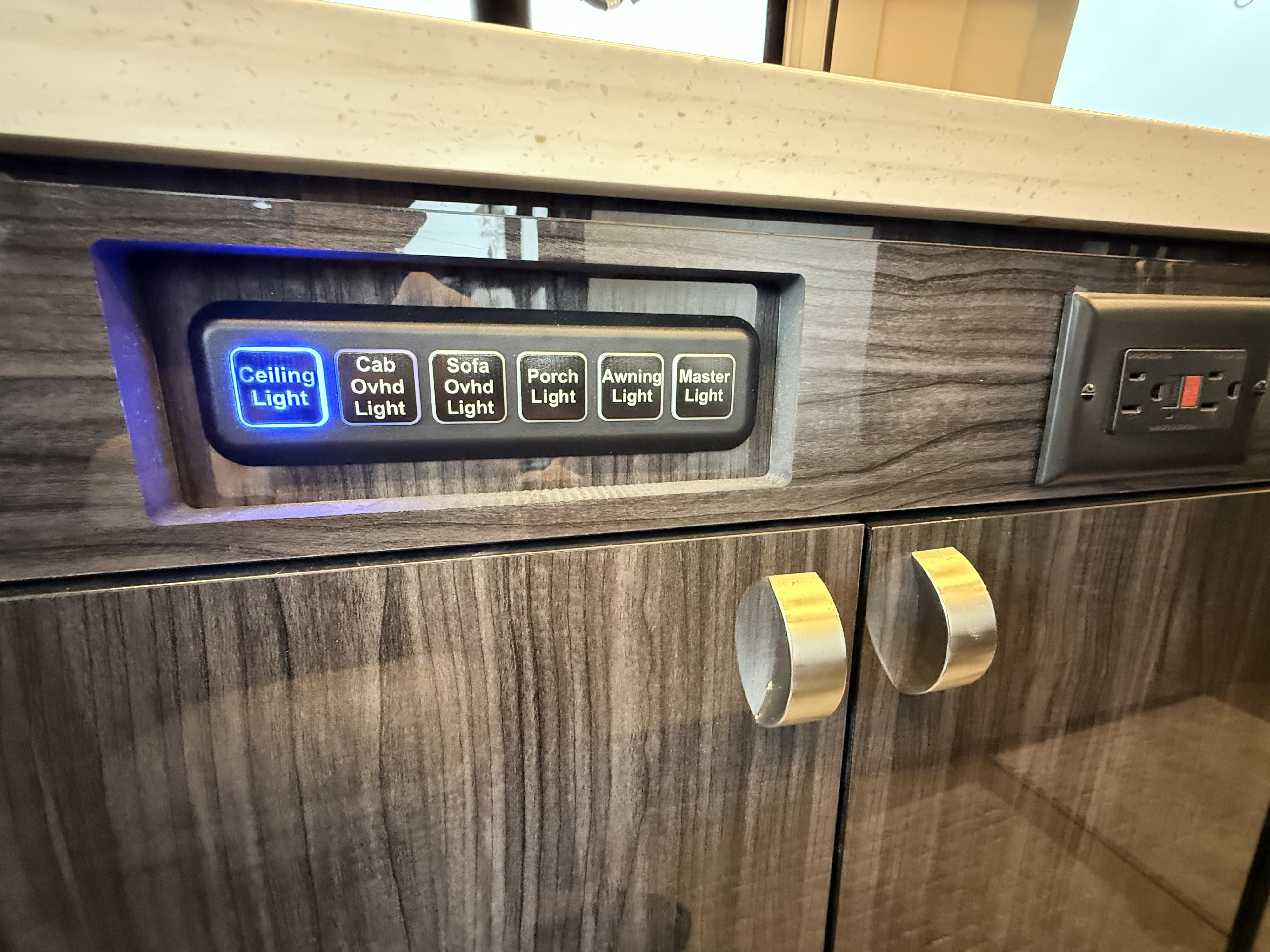 RV rental interior: illuminated light control panel (Ceiling Light on), electrical outlet, and wood-grained cabinetry.. Winnebago Era 2020