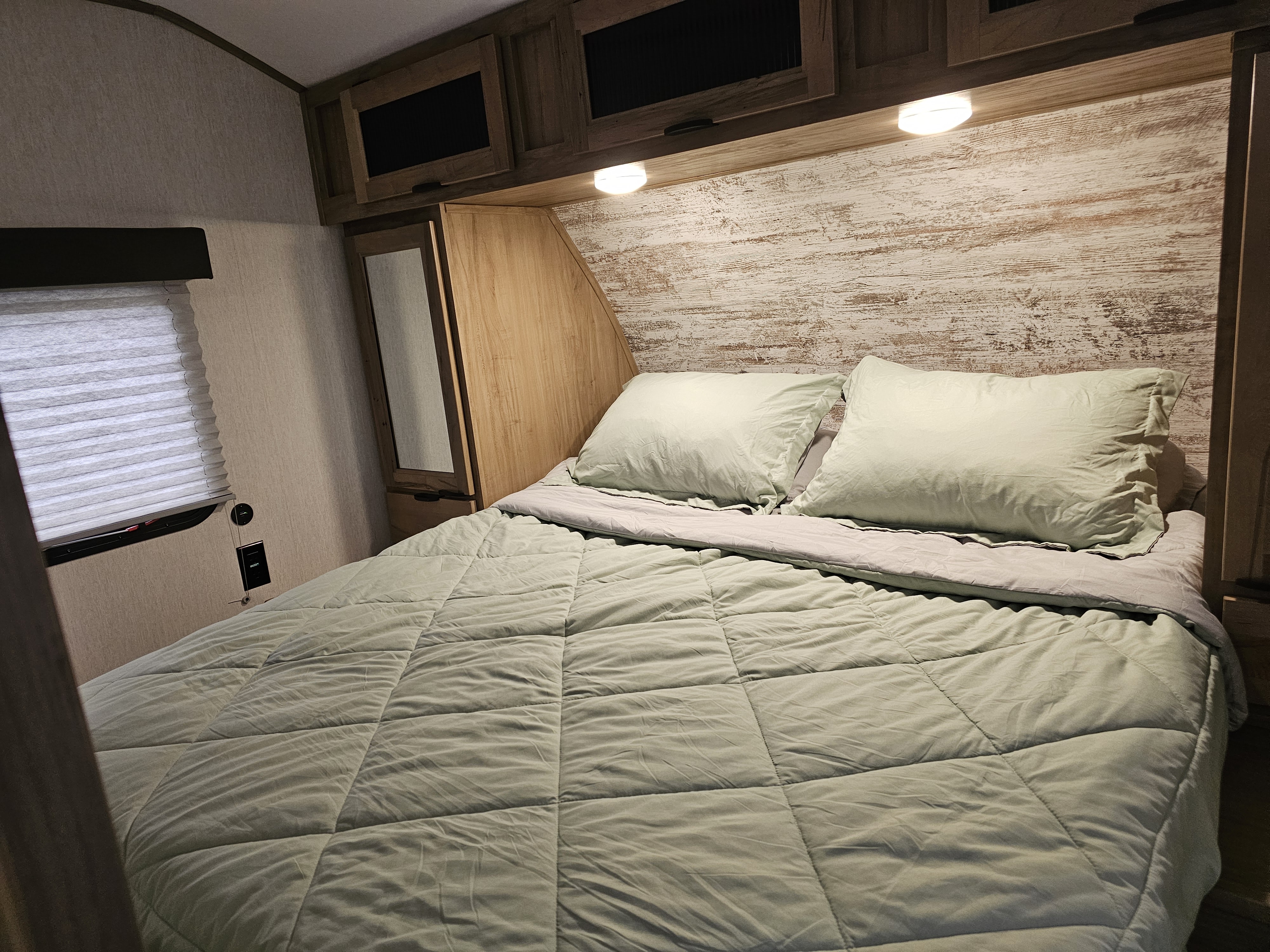 Comfortable RV rental bedroom with sage green bedding, distressed wood headboard, overhead cabinets, and window with blinds.. Forest River Cherokee Alpha Wolf 2024