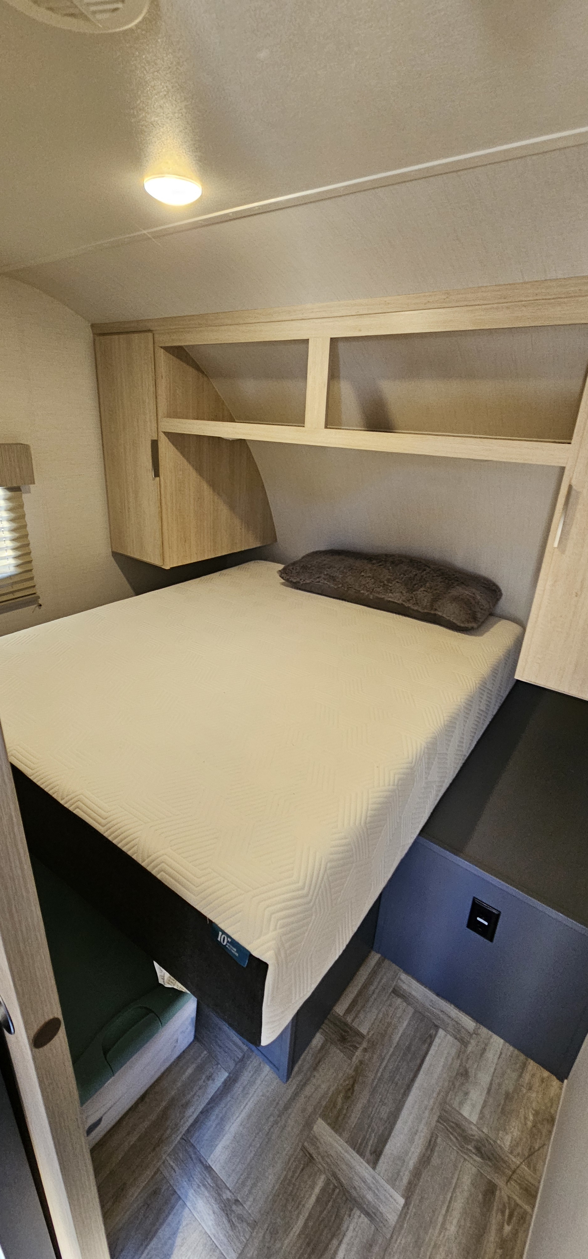 RV rental bedroom with bed, brown pillow, overhead wooden cabinets, and wood-look flooring. Light on ceiling.. Prime Time Tracer 2024