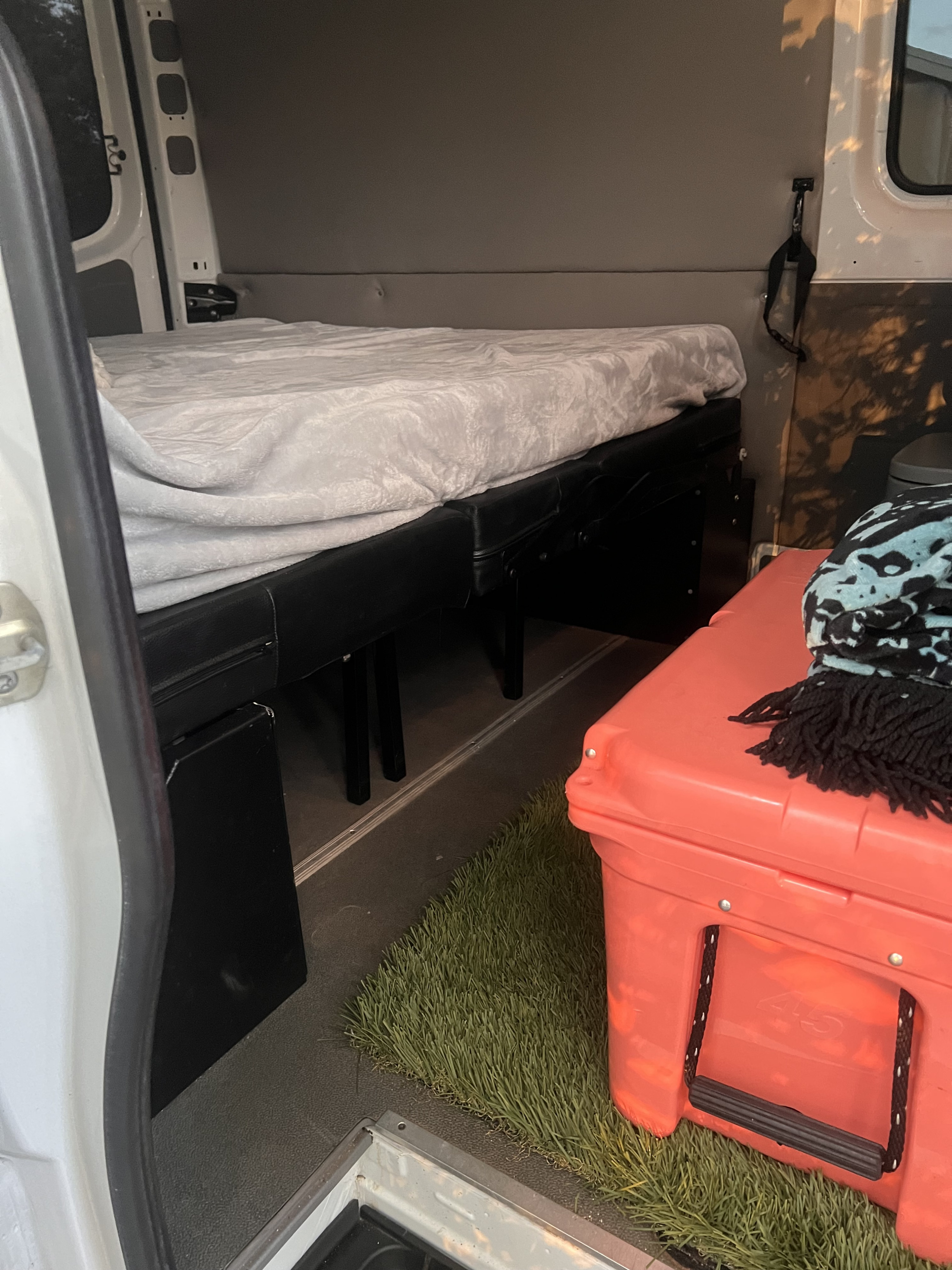 RV rental interior: A made bed with a grey blanket, bright orange cooler on a green turf mat, viewed from the open door.. Mercedes-Benz Sprinter 2017