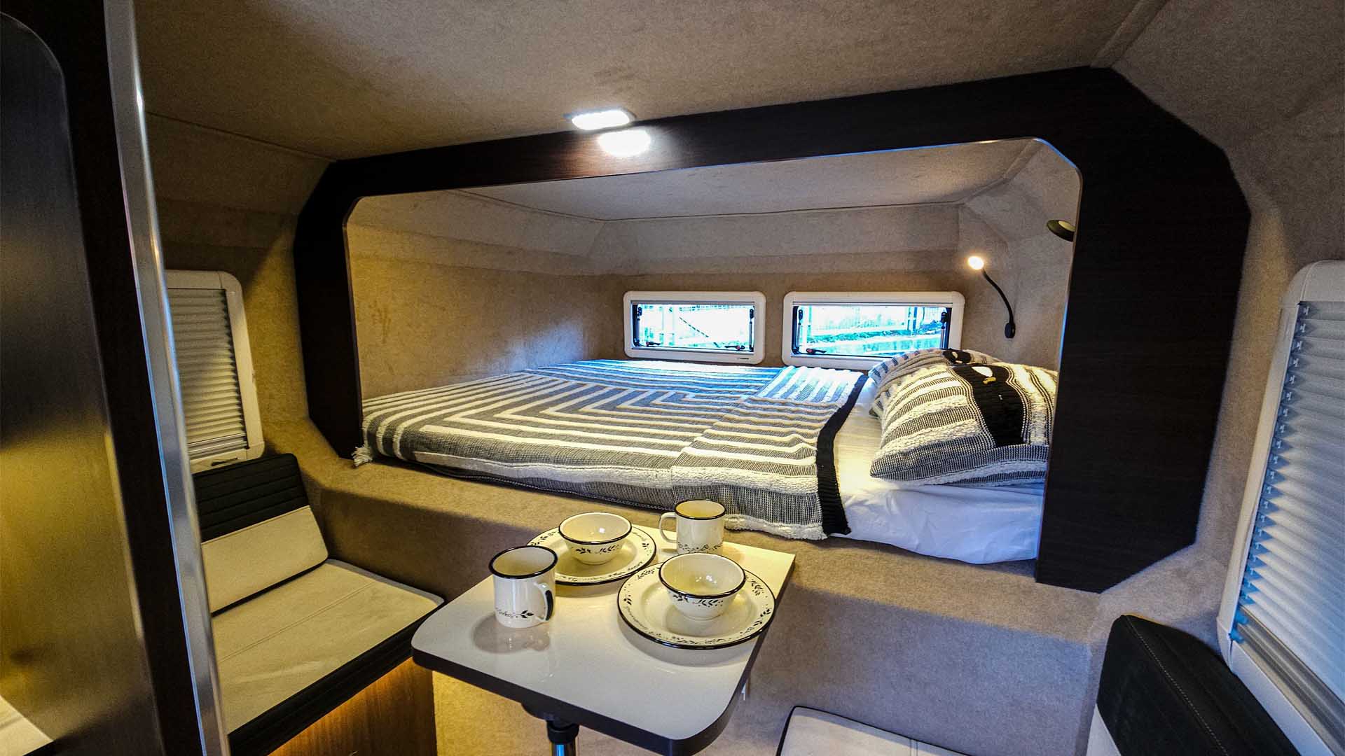Cozy rv rental interior: compact bedroom featuring a striped bed and windows, plus an adjacent dining area.. F-150 Lightning / Hotomobil Gladiator XL with roof tent 2023