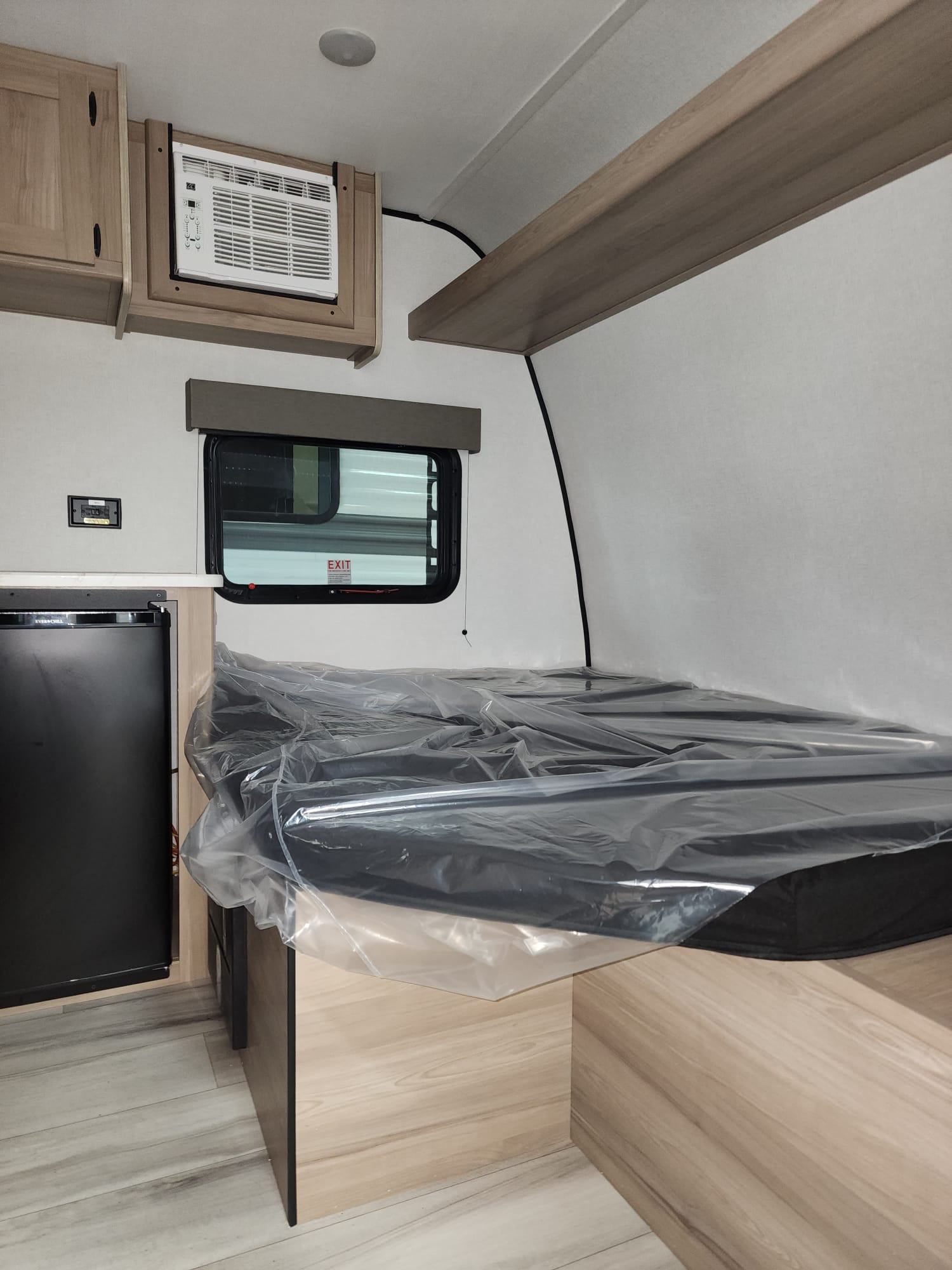 Interior of an rv rental showing a compact fridge, window AC unit, and a new bed still wrapped in protective plastic.. Keystone Coleman 13B 2025