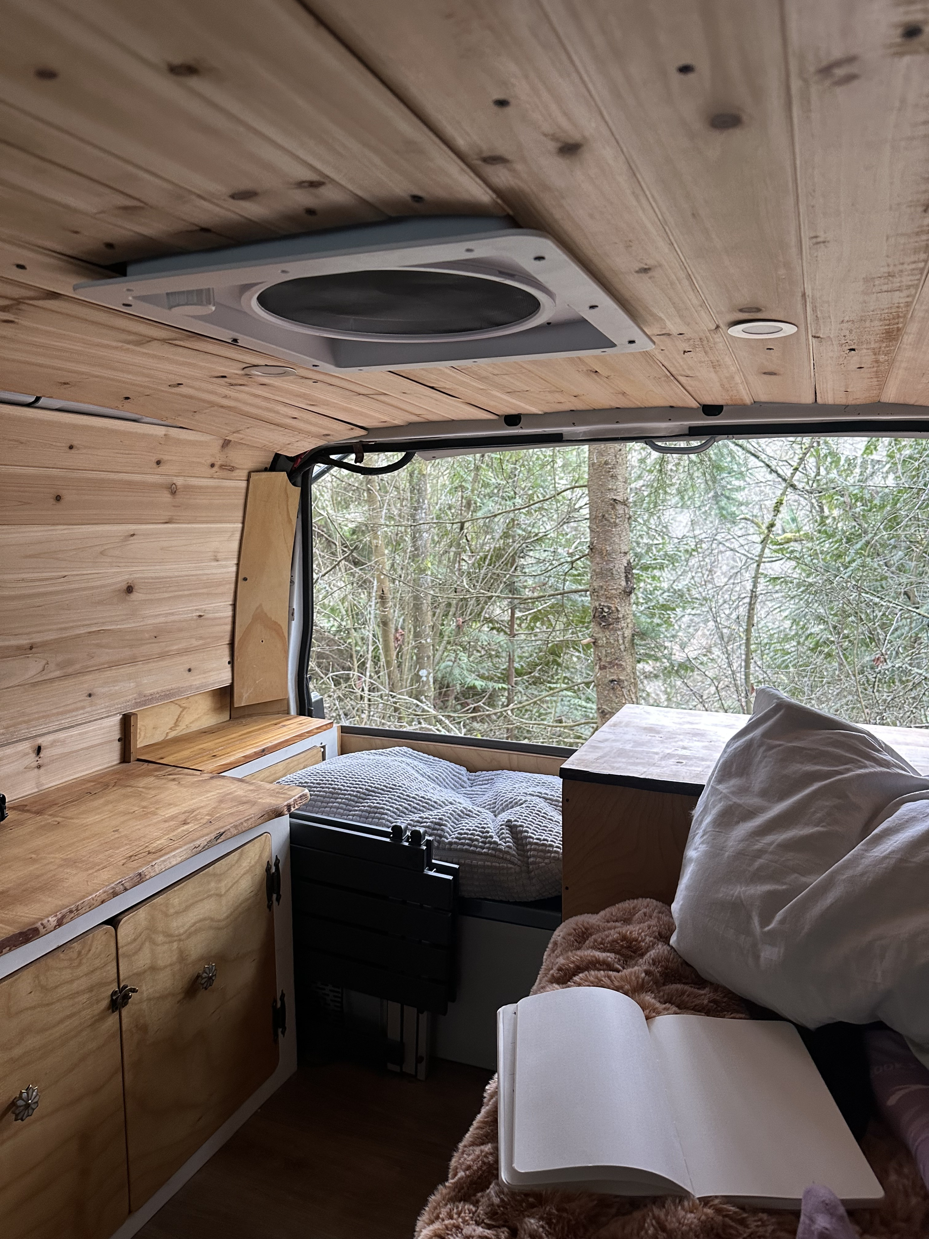 Cozy camper van RV rental interior with wooden walls & ceiling. Bed, kitchen area, & open notebook overlook a wintry forest view.. Chevrolet Astro 2003